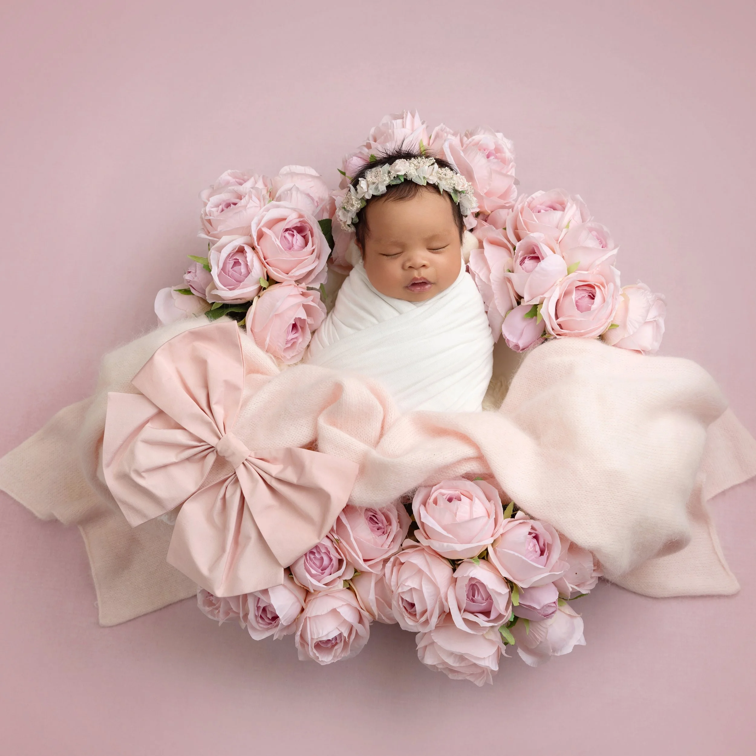A newborn baby wrapped in a white blanket, lying on a pink background, surrounded by pink roses, wearing a floral headband, with a pink bow and cream-colored blanket.