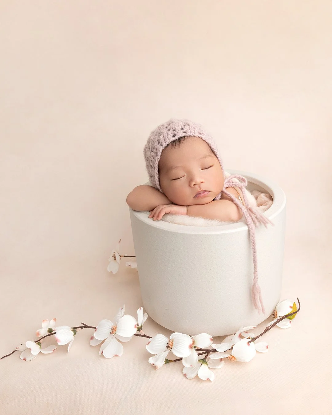 A sleeping baby wearing a pink knit hat with a string, resting on their arms inside a white cylinder-shaped container, surrounded by white flowers on a neutral background.