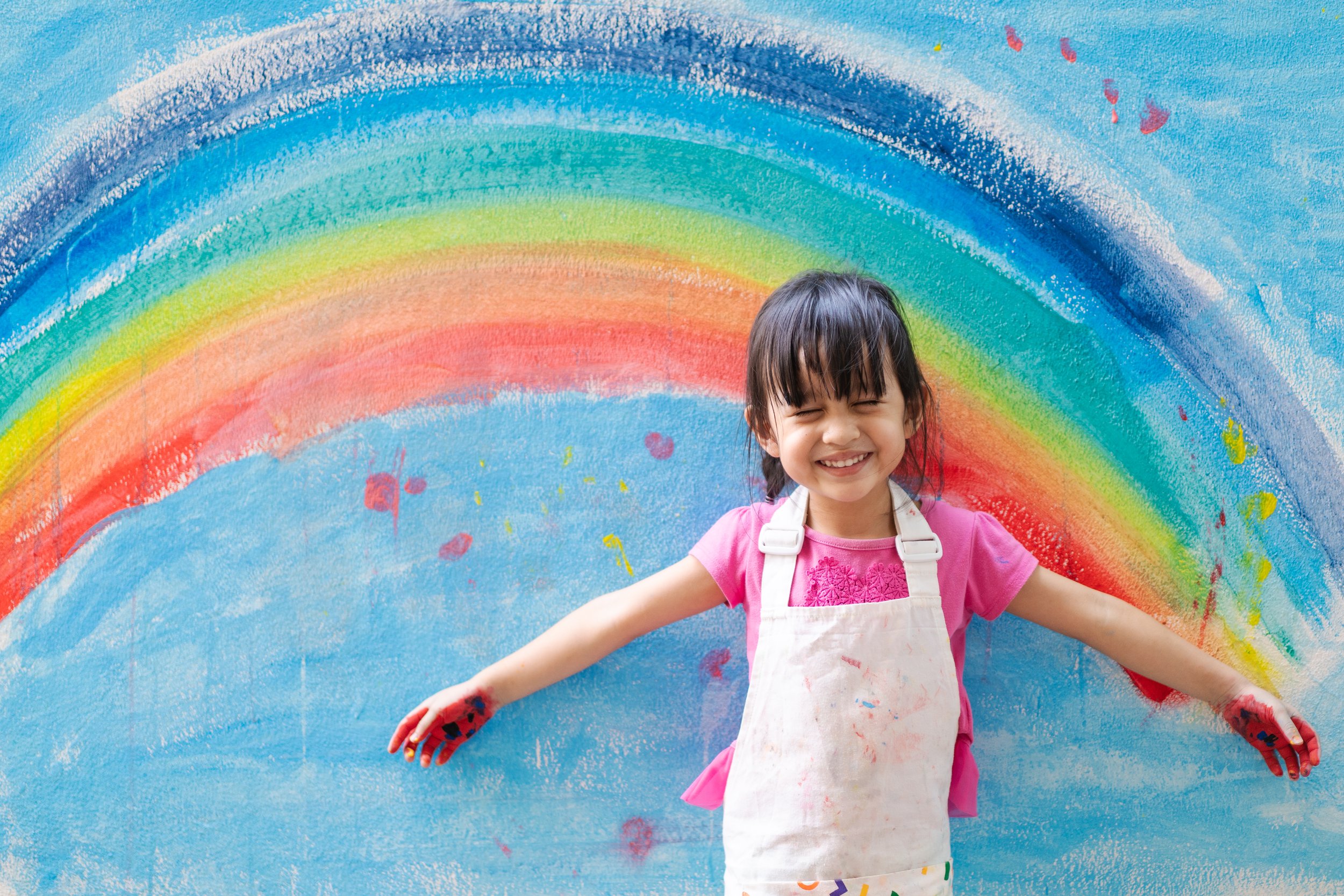 A child stands in front of a rainbow mural