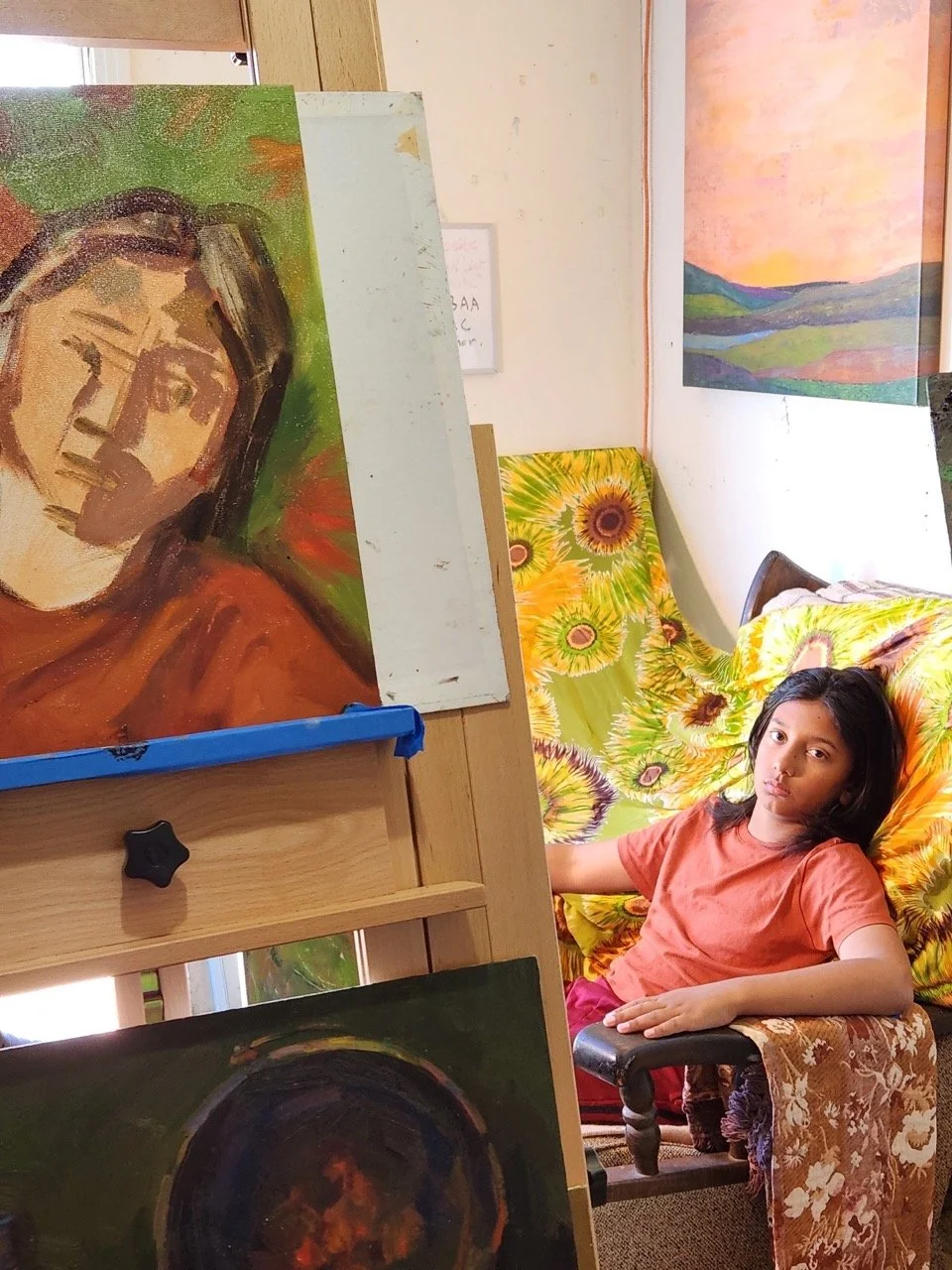 A young boy relaxing on a chair with a floral blanket, surrounded by colorful paintings including a landscape and sunflower paintings, in an art studio or gallery.