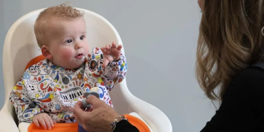 A child sits in a high chair and reaches out with their hand toward a spoon of food.