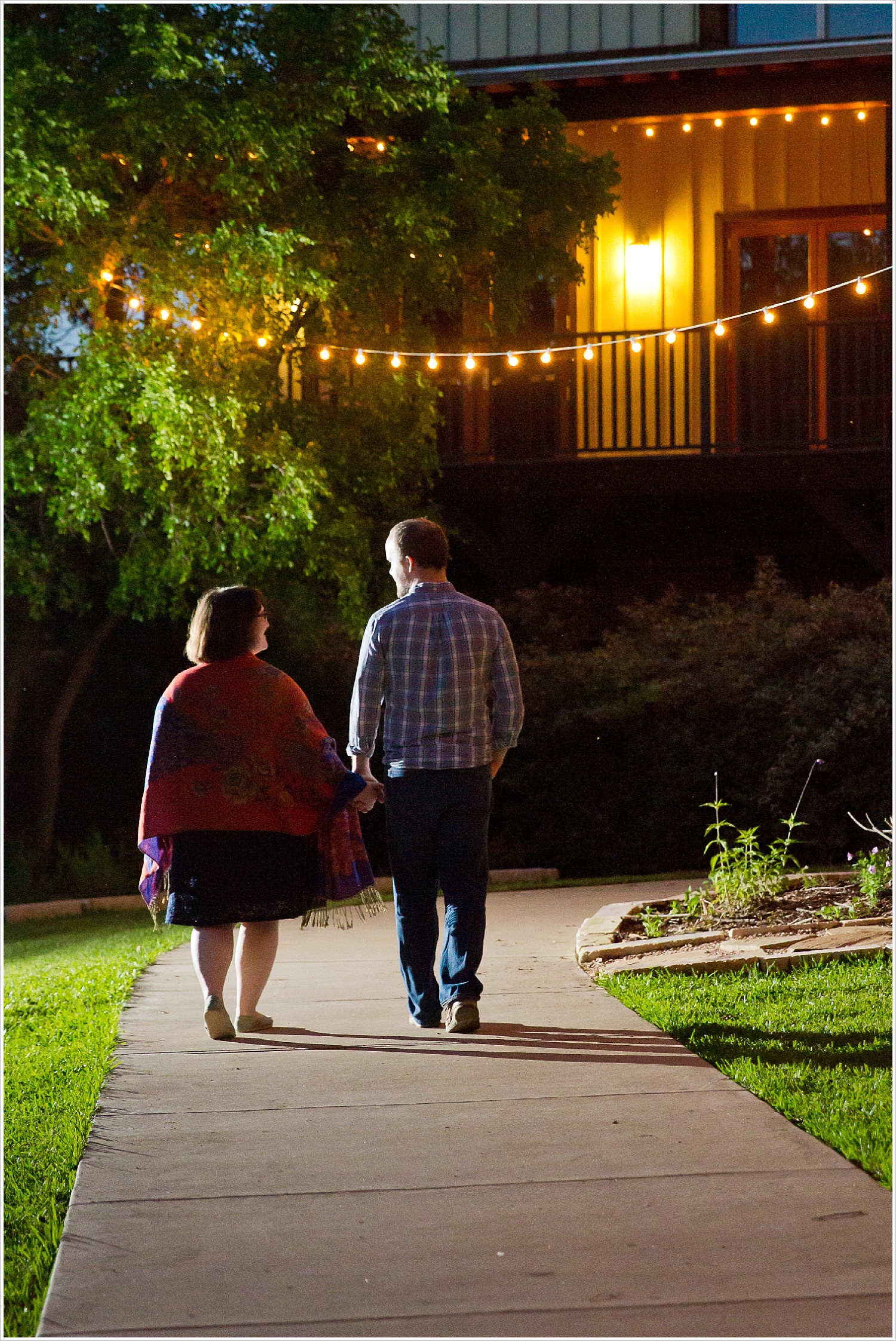 Couple walks away from camera, hand-in-hand, under string lights at Carleen Bright Arboretum in Woodway, Texas