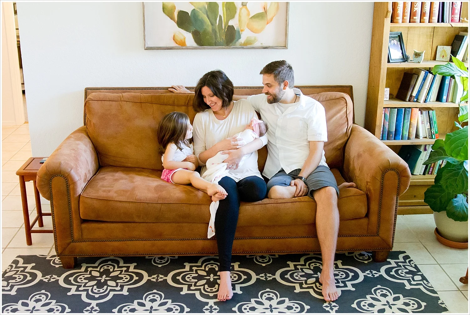 A mom and dad hold their newborn daughter while little sister looks on in their living room during a lifestyle in-home portrait session in Waco, Texas - Jason & Melaina Photography - www.jasonandmelaina.com