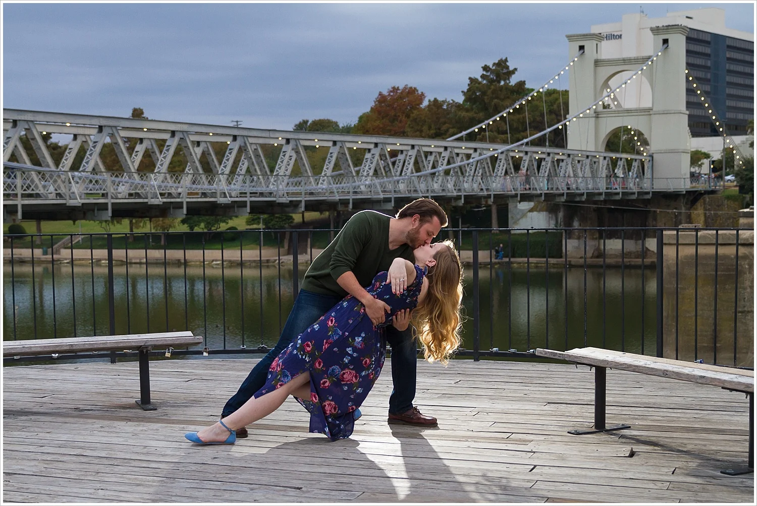 A groom dips his fiance and kisses her during their engagement portrait session near the Suspension Bridge in Waco, Texas - Jason & Melaina Photography - www.jasonandmelaina.com