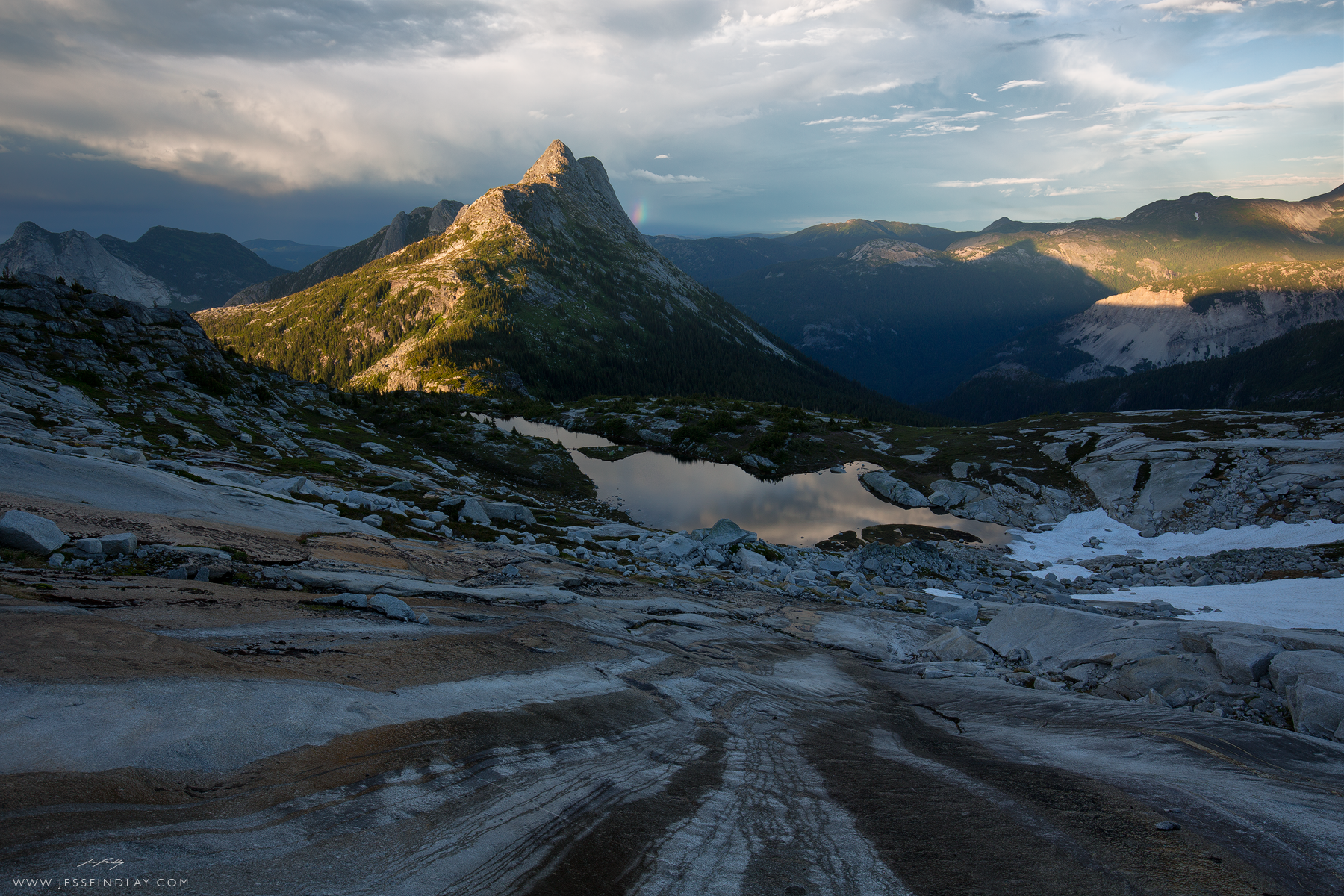 Dramatic sunset skies loom over the Cascade Mountains of southwestern British Columbia, where snow melt trickles down towards an alpine tarn. My tent can barely be seen on a small island below.