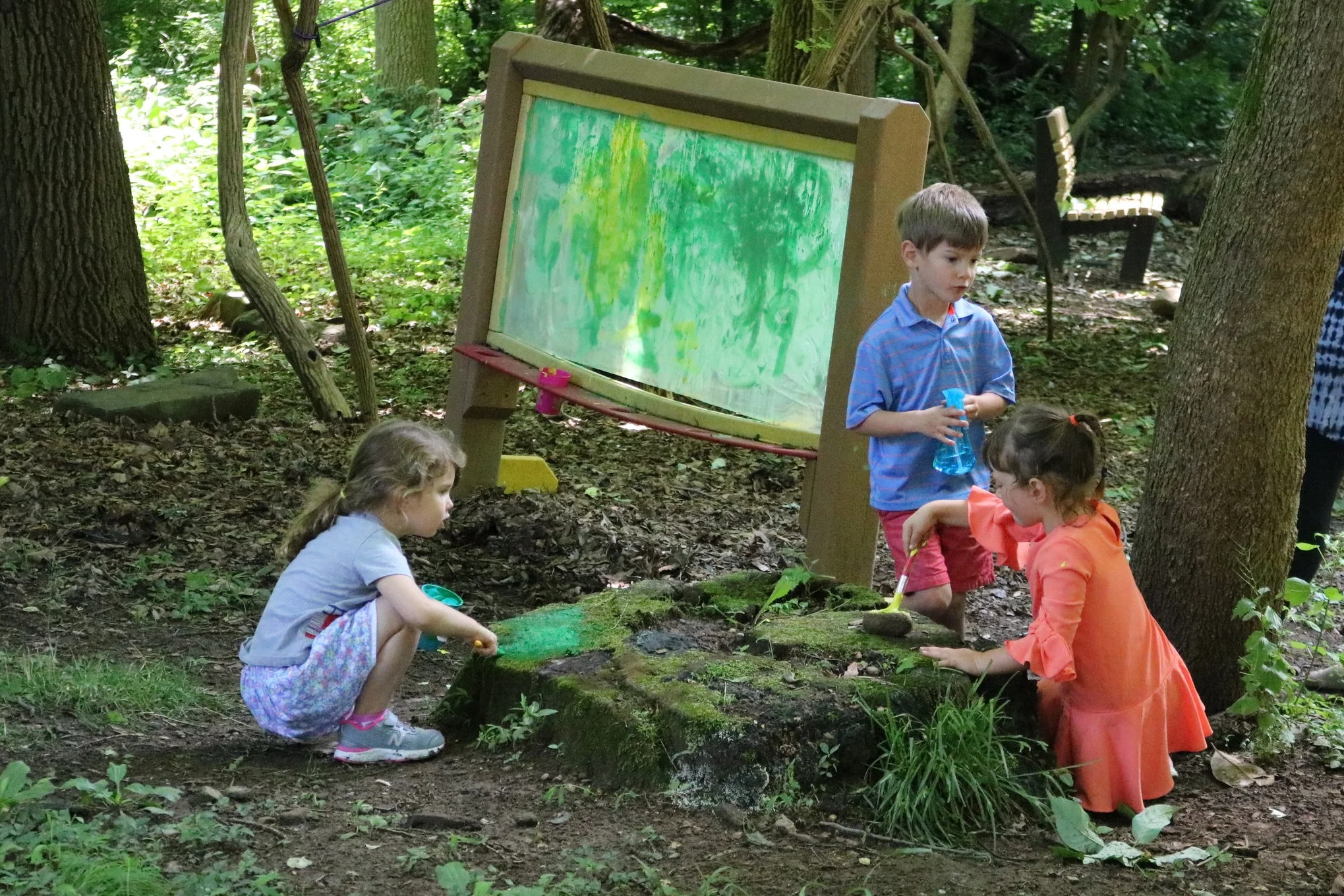 Three preschoolers playing outside, painting rocks in forest