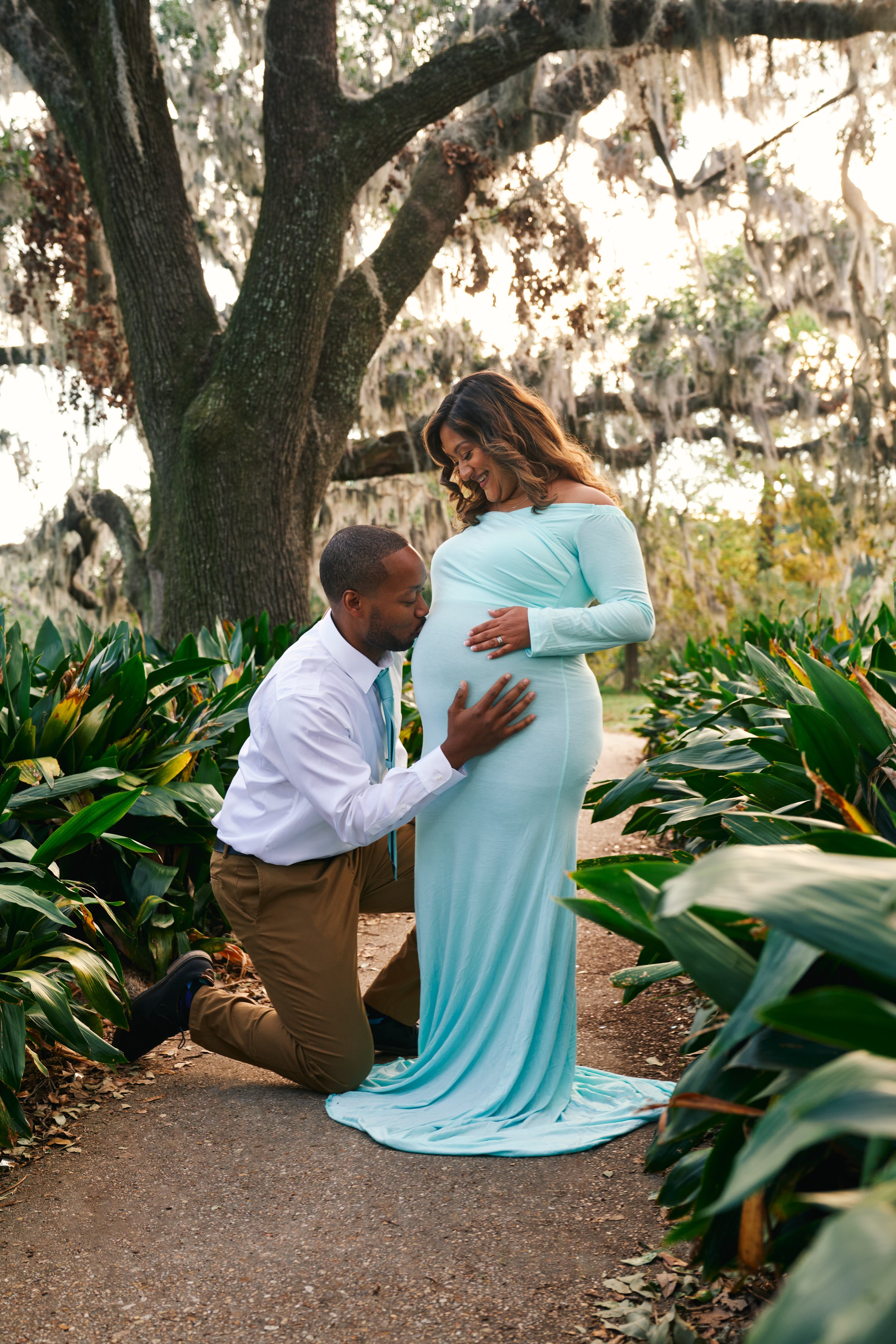 A man kneeling and holding a pregnant woman's belly, smiling at her, in a garden with green plants and a large tree in the background during sunset.