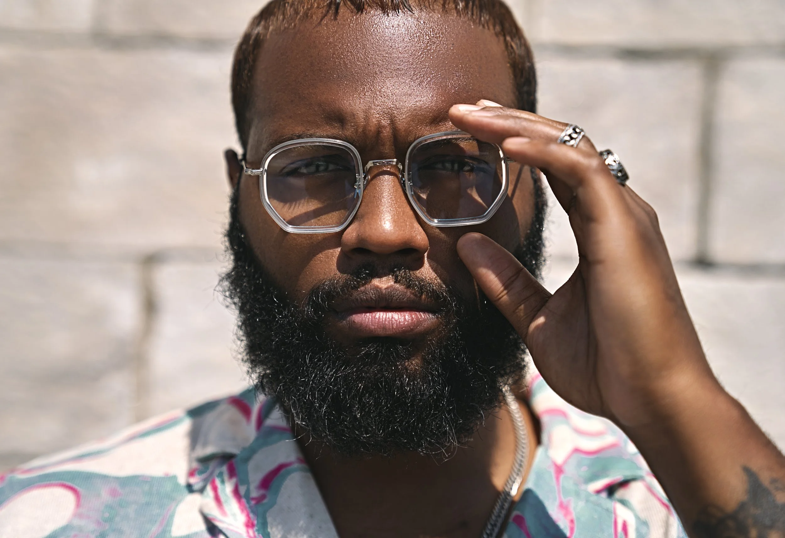 Close-up of a man with a beard wearing glasses and a striped shirt, holding his sunglasses with his right hand, outdoors on a sunny day.
