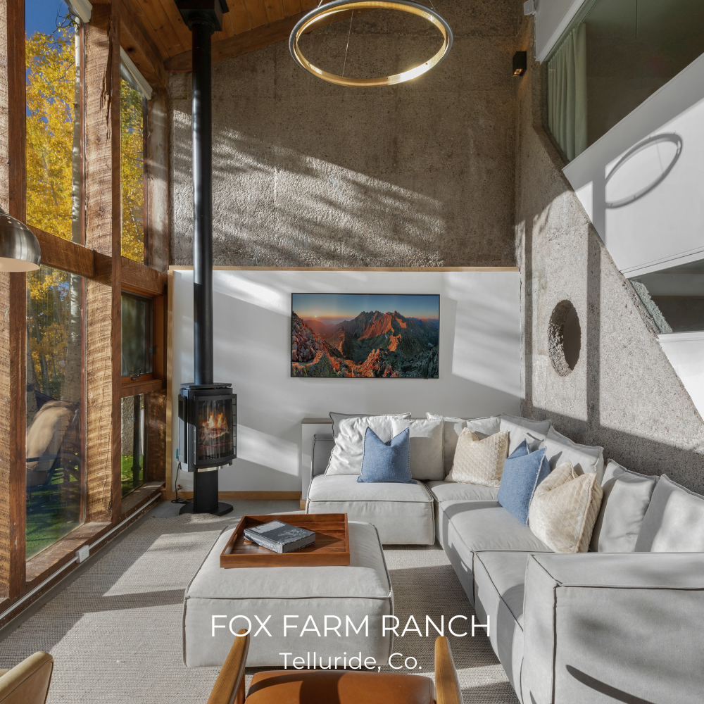 Modern living room with large windows, gray sectional sofa, blue and cream pillows, a wall-mounted fireplace, and nature photograph. The space is within Fox Farm Ranch, Telluride, Colorado.