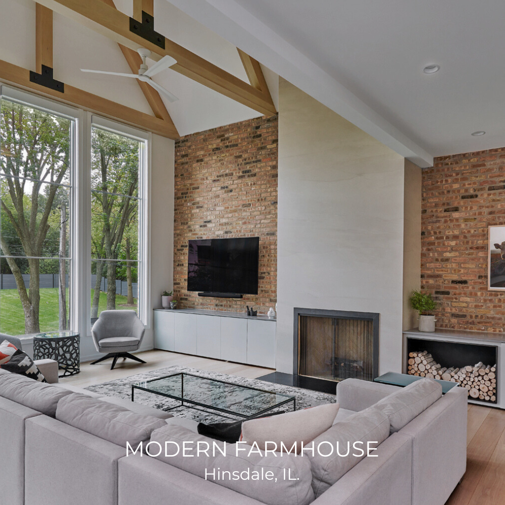 Modern farmhouse living room interior in Hinsdale, IL with large windows, exposed brick wall, ceiling fan, wall-mounted TV, minimalist furniture, and a fireplace.