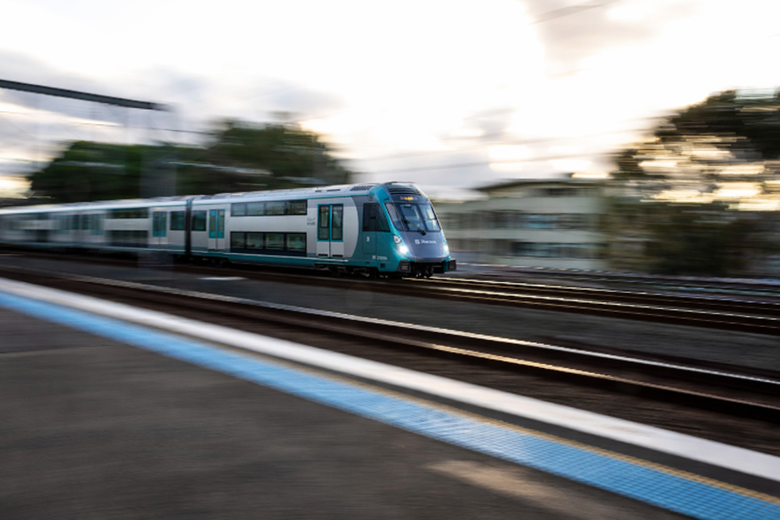A modern blue and white train moving quickly on railway tracks during sunset with blurred background and motion blur.