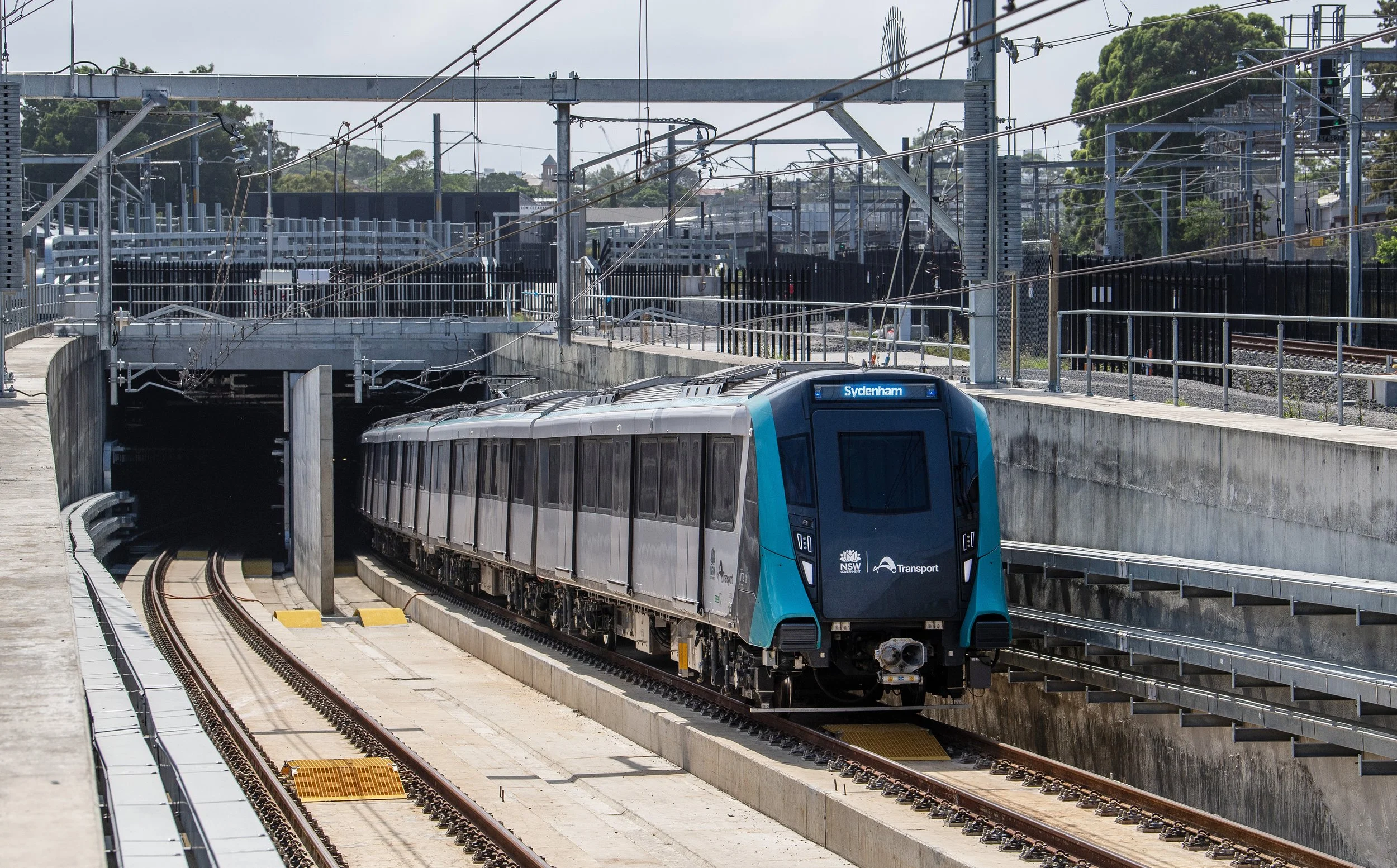 A modern light blue and gray train traveling through a train station tunnel beneath a city street, with overhead electrical wires and infrastructure.