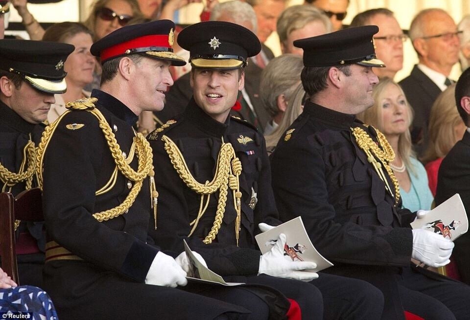 Three men in formal military dress uniforms with gold braid and insignia, sitting in a row at a formal event, surrounded by an audience of mostly older individuals.
