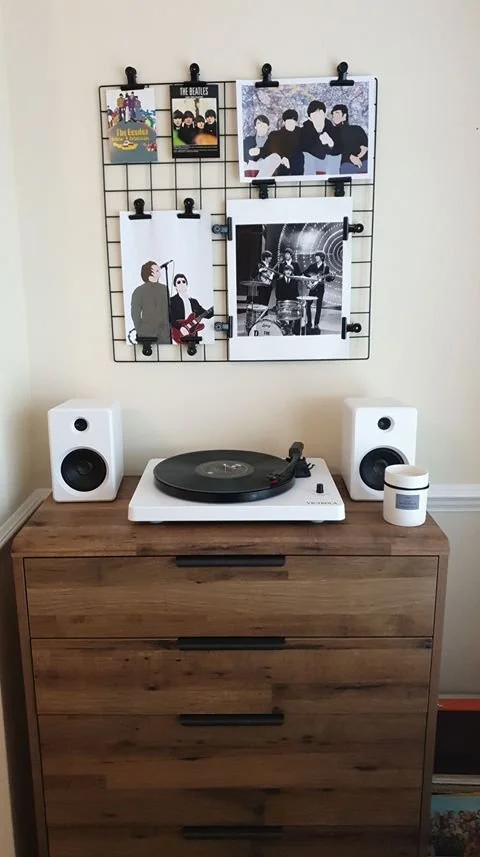 A record player on a wooden dresser with two white speakers, a small white container, and wall mounted photos of The Beatles and other musicians.