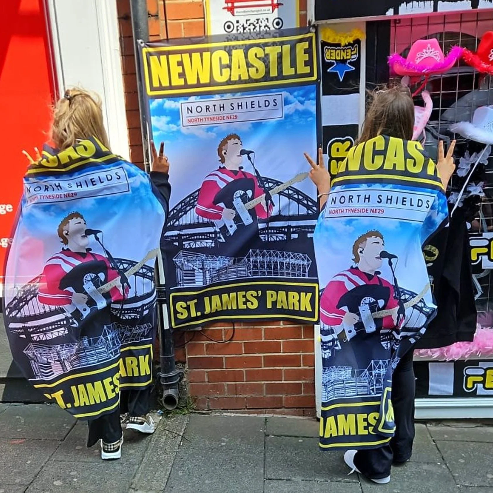 Two children standing outside a brick building, facing a large Newcastle United football club banner on the wall. The children wear black clothing and Newcastle United scarves and jackets with the team name and a stadium illustration. They hold up pe