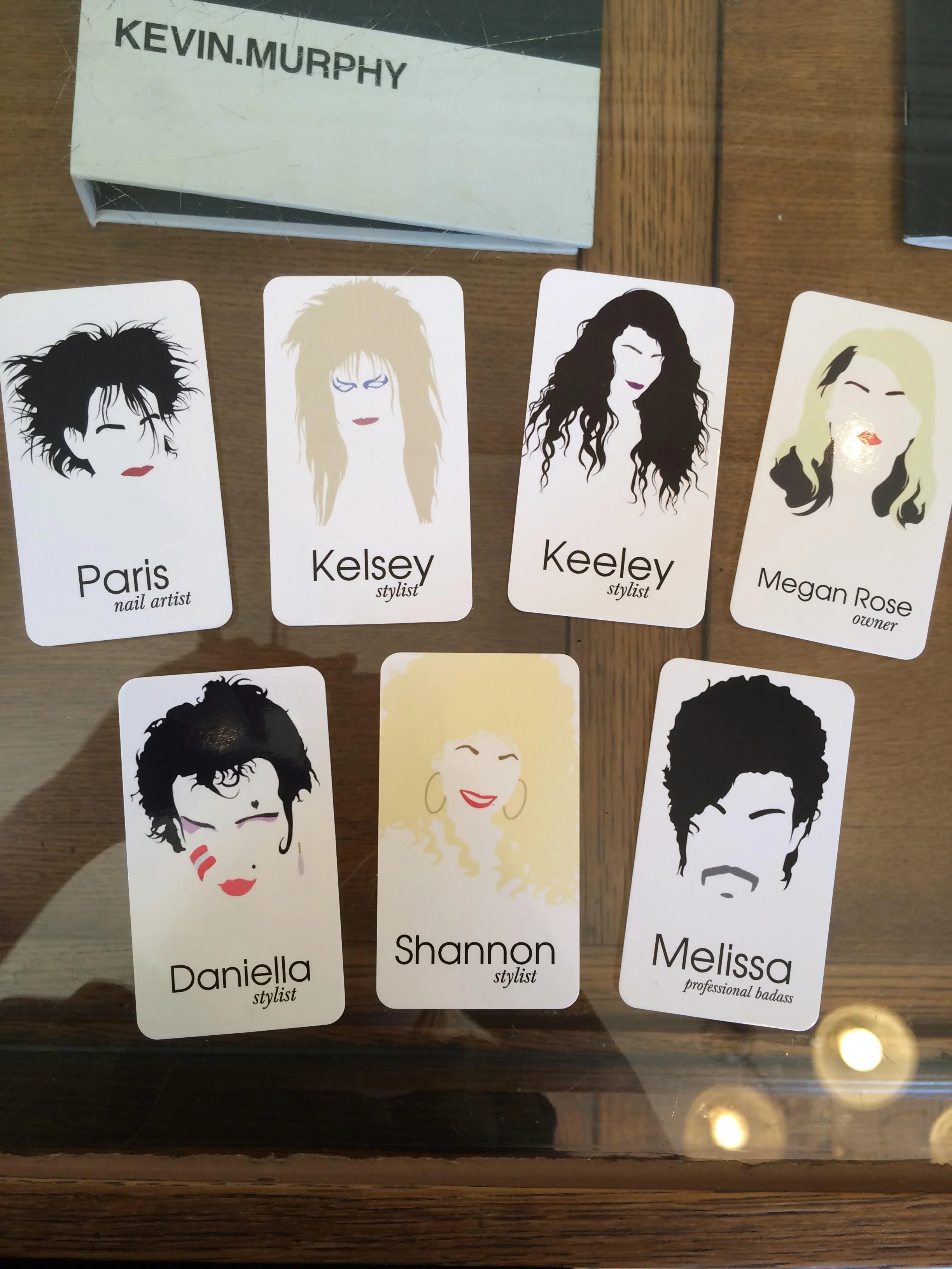 A collection of illustrated portrait cards representing different women with various hairstyles and makeup, each labeled with a name and styling role, placed on a wooden surface.