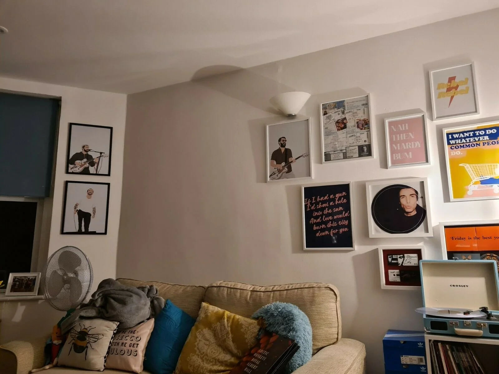 Living room wall decorated with framed posters and artwork, including images of musicians and quotes, above a sofa with multicolored pillows. A small fan and boxes are visible on a side table and the floor.