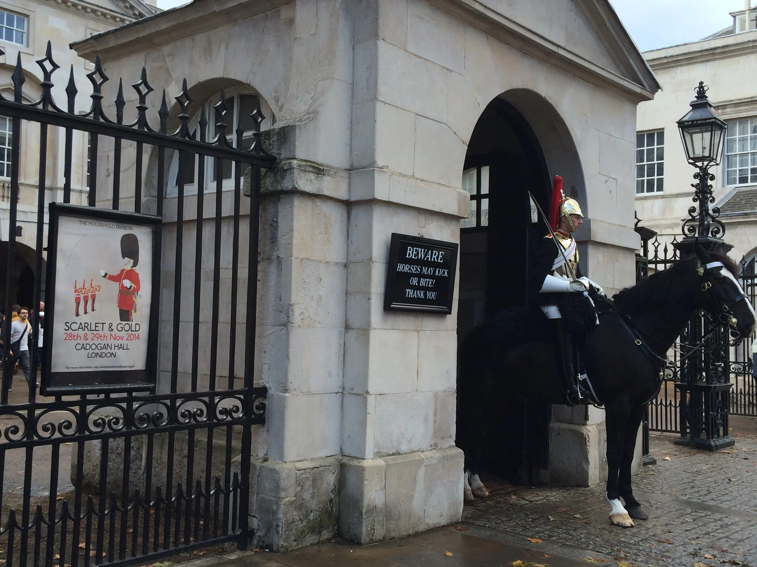 A guard in traditional uniform on horseback standing in front of a gate with a sign warning about the horses kicking or biting, near a stone building with large windows at Cadogan Hall in London.