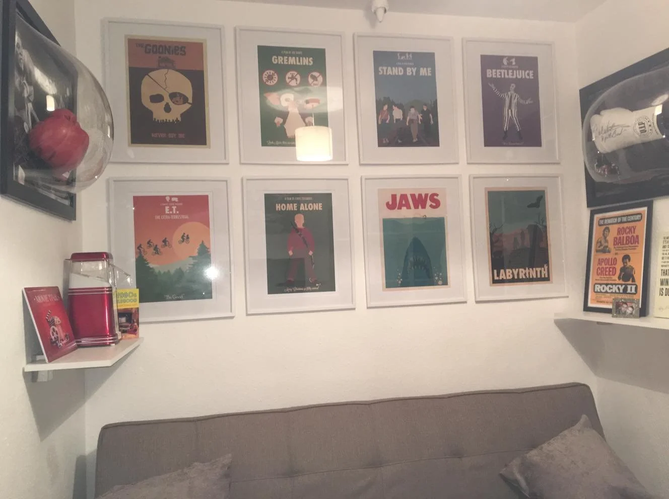 A bedroom wall decorated with nine framed movie posters arranged in a grid, featuring titles like 'The Goonies,' 'Gremlins,' 'Stand by Me,' 'Beetlejuice,' 'E.T.,' 'Home Alone,' 'Jaws,' and 'Labyrinth.' There are wall-mounted shelves on each side of t
