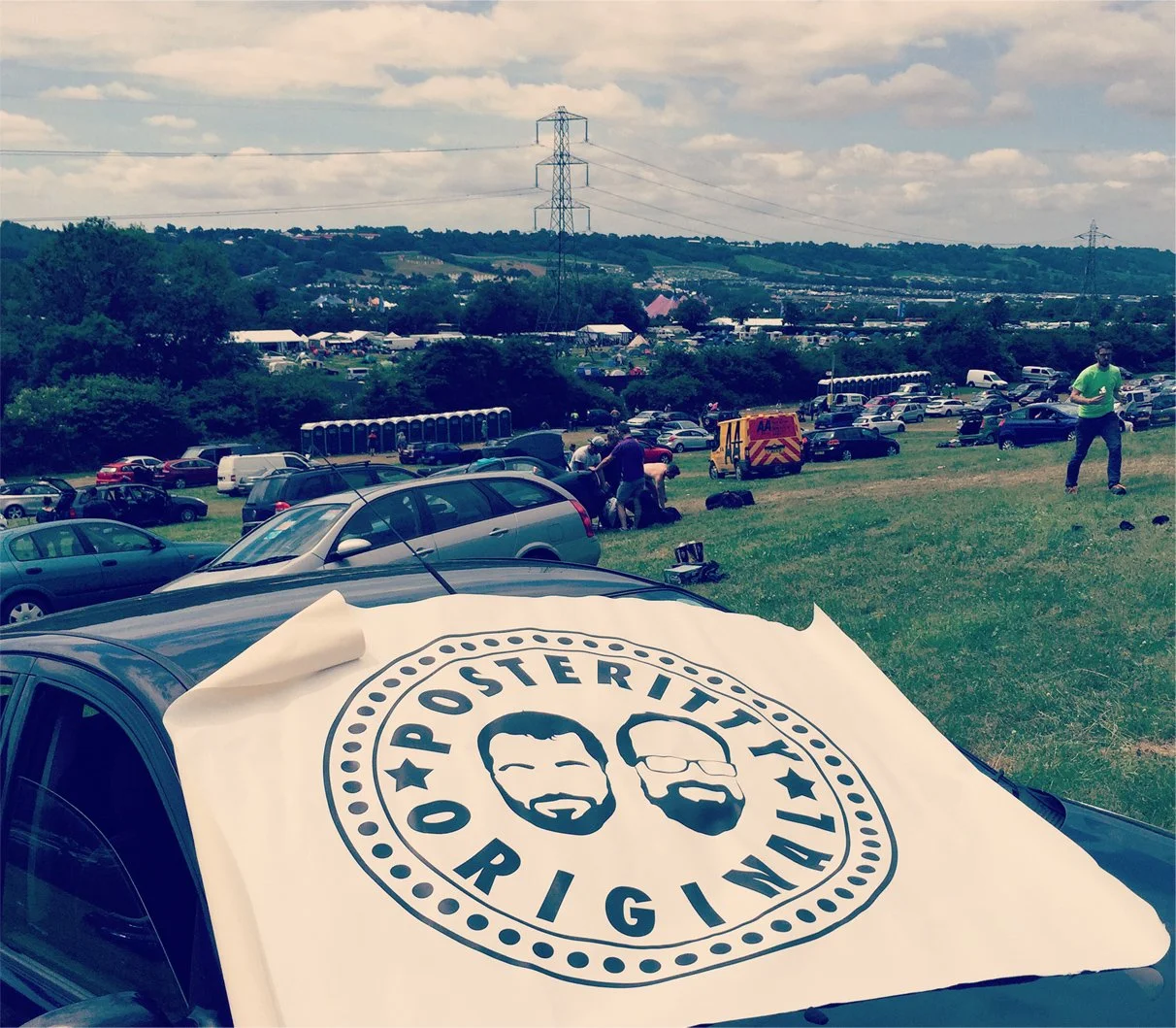 An outdoor event with many parked cars on a grassy hill, people gathered in small groups, and a large poster on a car with the logo 'Posterity Original' featuring two men's cartoon faces.