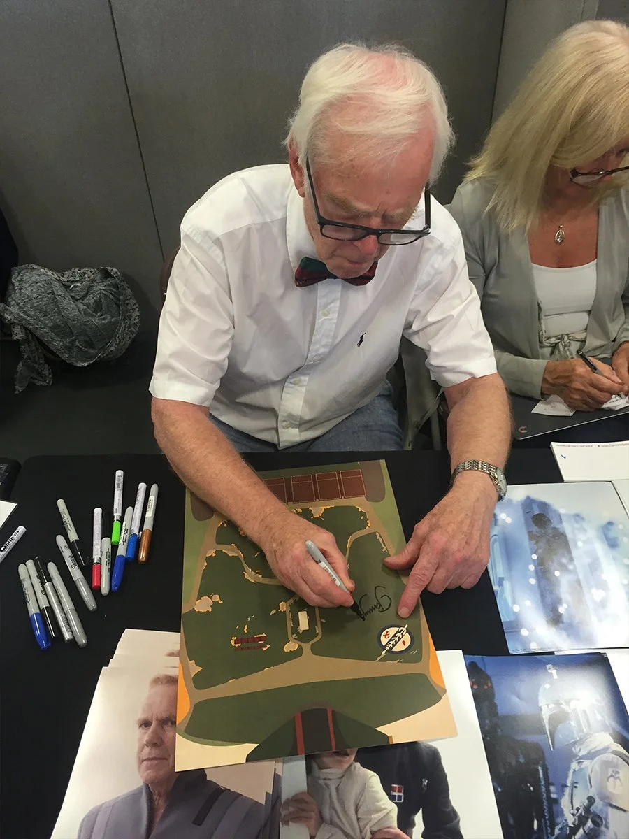 An elderly man with white hair, glasses, and a bow tie is sitting at a table, signing an illustrated map. Next to him, a woman with blonde hair and glasses is also seated, writing on a notepad.