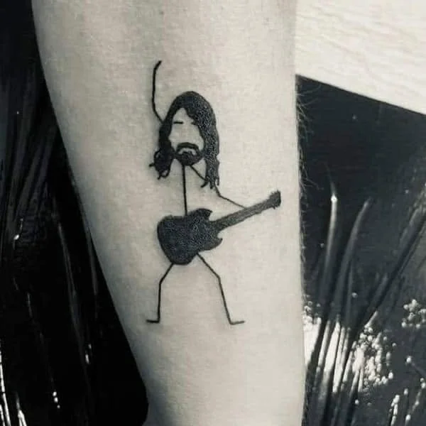 Tattoo of a stick figure with long hair and beard playing an acoustic guitar.