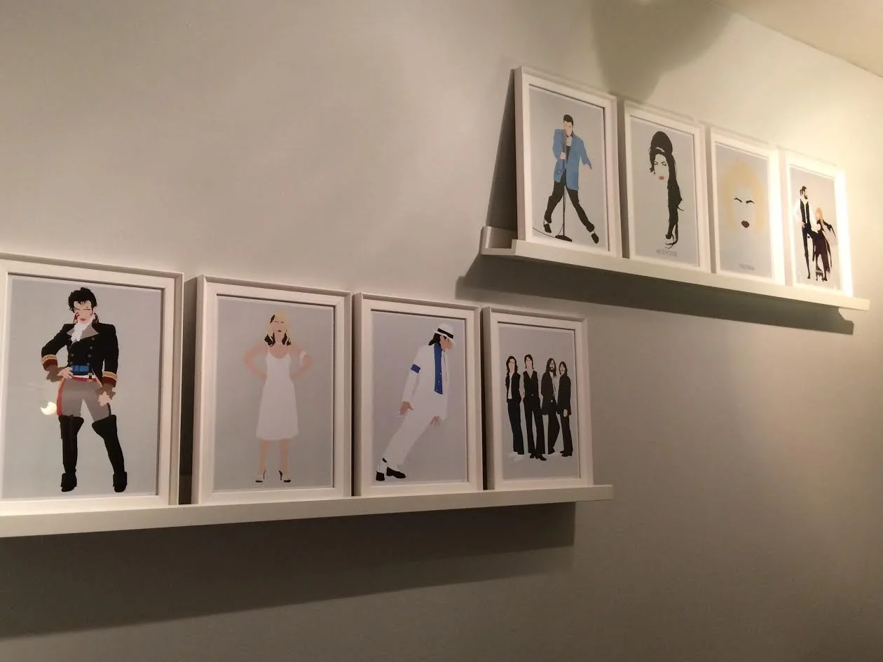 Collection of nine framed fashion illustrations displayed on two white shelves on a plain wall, depicting various stylish women and men in different outfits and poses.