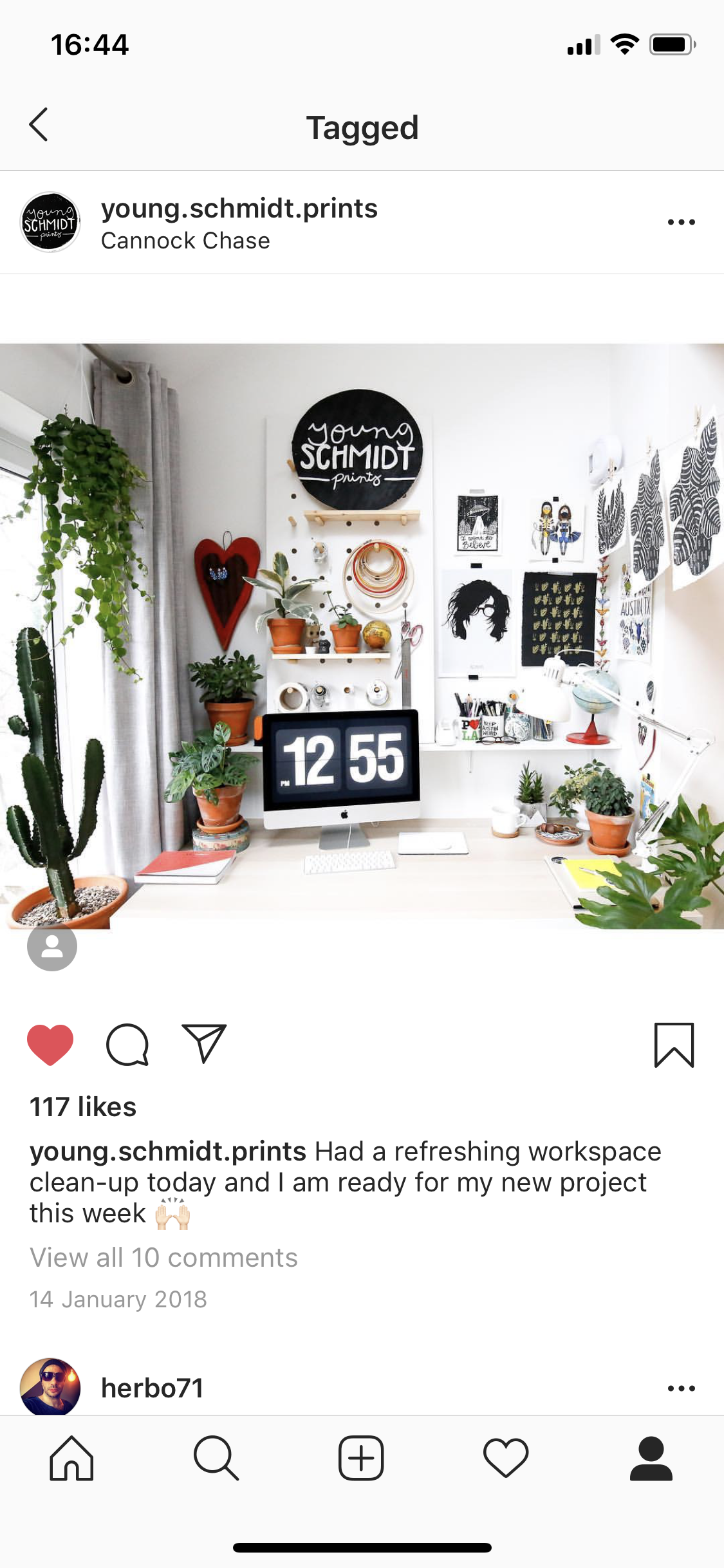 Bright workspace with a white desk, an iMac showing the time 12:55, surrounded by plants, art, and office supplies. Decor includes black and white wall art, shelves with small potted plants, a cactus, and a large circular sign reading 'Young Schmidt 