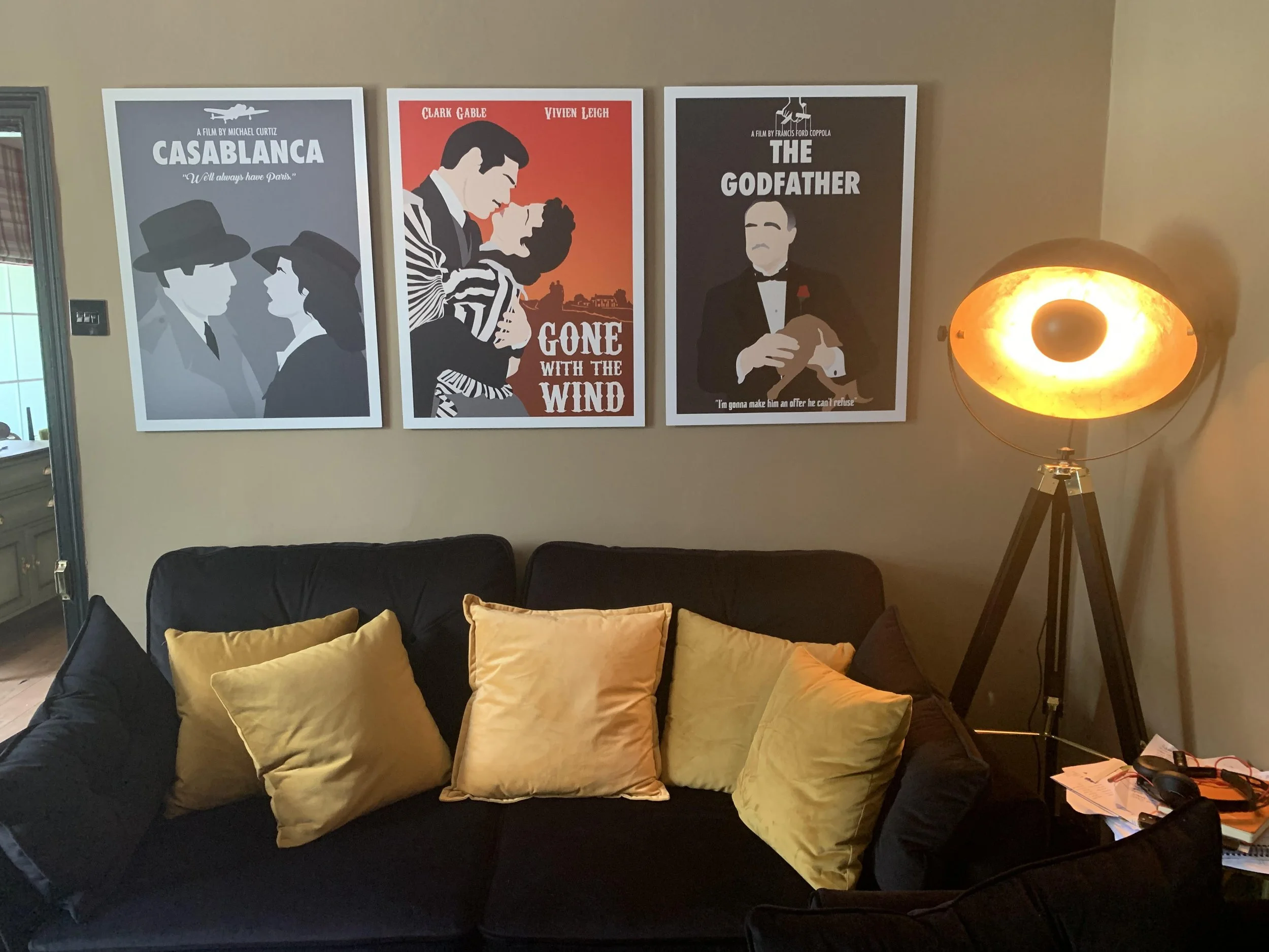 Living room wall decorated with three movie posters from Casablanca, Gone with the Wind, and The Godfather. A lamp with a glowing orange eye shape is standing next to the posters. A black sofa with multiple yellow pillows is in front of the wall, and