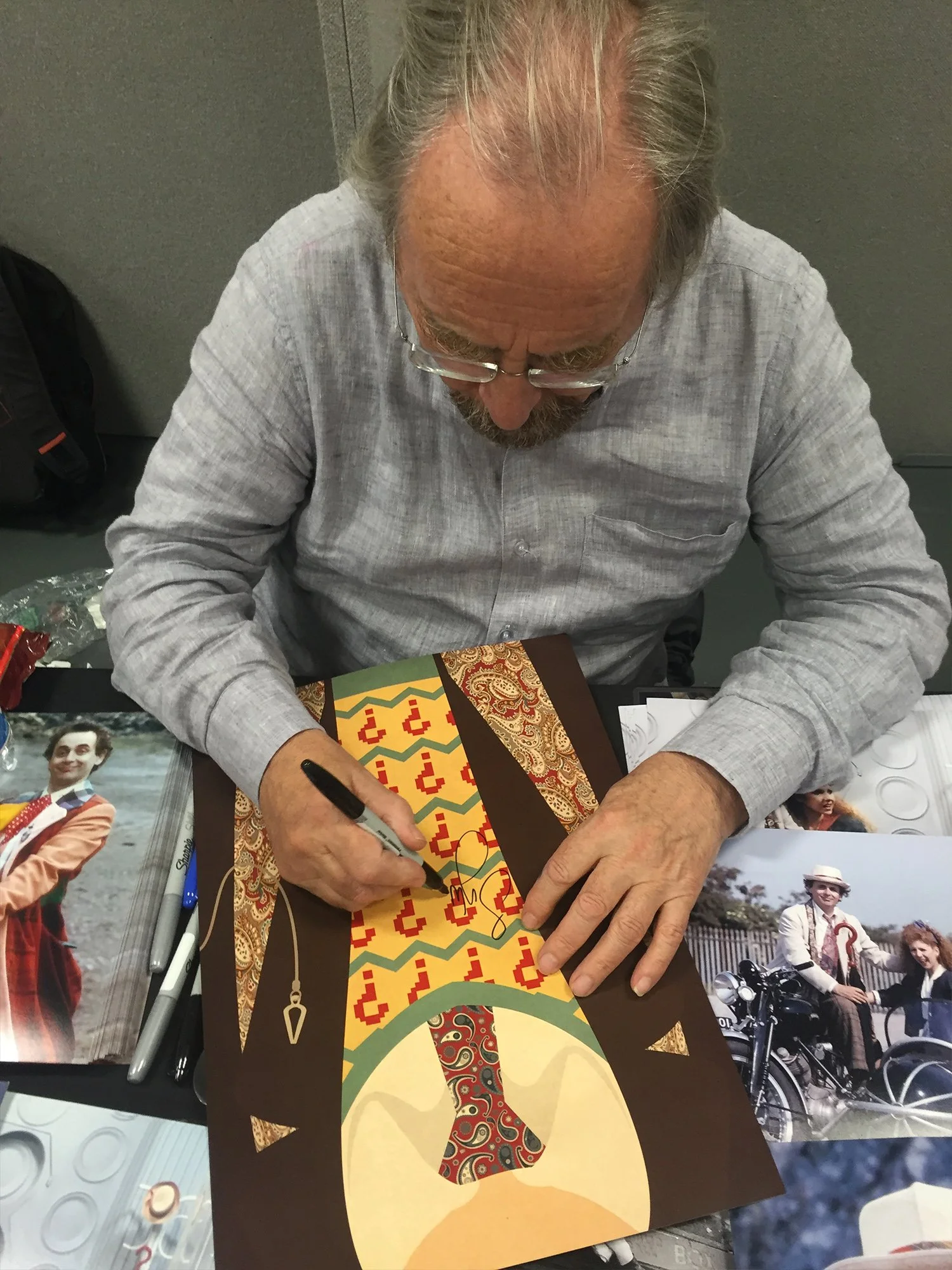An elderly man with glasses is signing a colorful artwork with a black marker. The artwork features a stylized figure with a patterned dress, set against a warm background. There are photos of people and motorcycles on the table, indicating a signing
