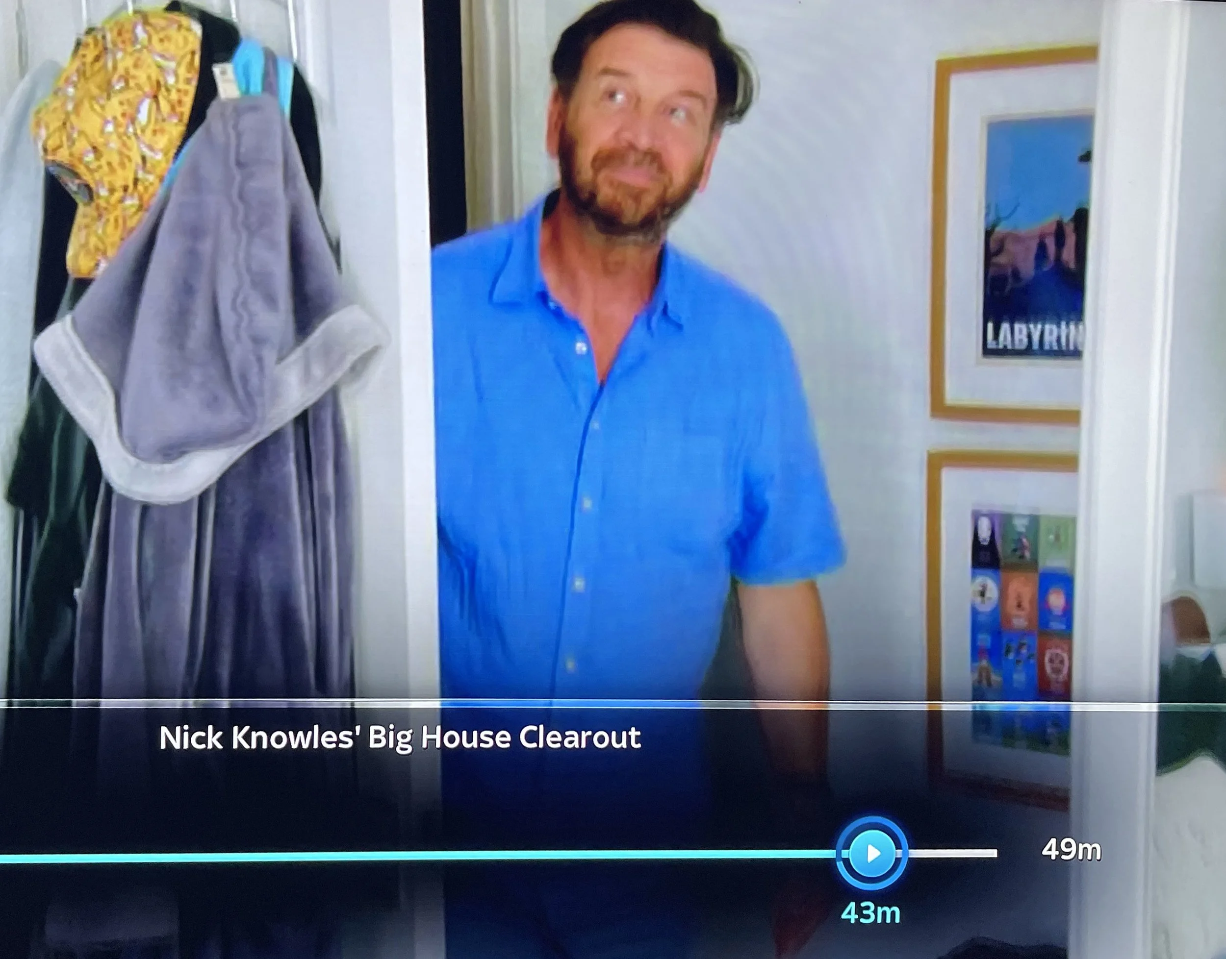 Partial view of a television screen showing a scene from the show Nick Knowles' Big House Clearout, with a man with dark hair and a beard wearing a blue shirt standing near a door, and a hallway with family pictures on the wall.