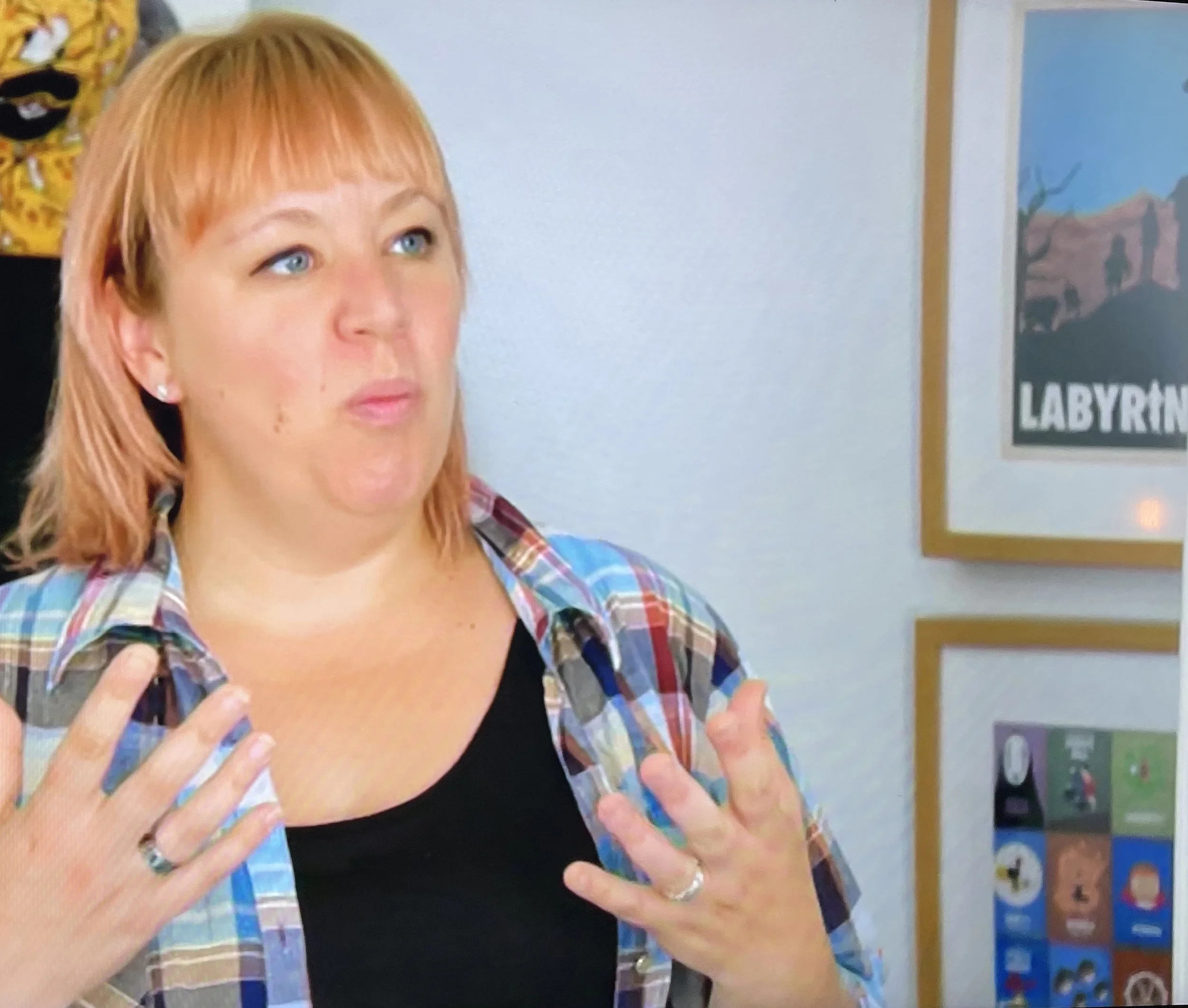 A woman with reddish hair, wearing a plaid shirt over a black top, gesturing with her hands as she speaks. In the background, there is a wall with colorful artwork and a sign that reads "LABYRINTH."