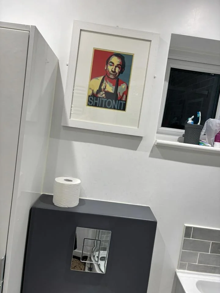 A framed pop art style portrait of a man with dark hair and a collared shirt, featuring the word "SHITONIT" at the bottom, hanging on a white wall in a bathroom or kitchen area.