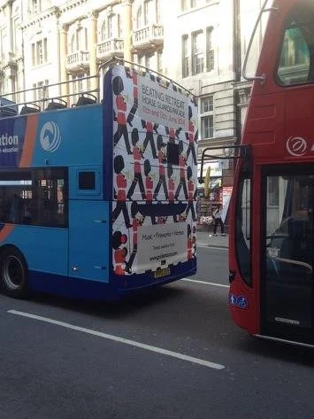 Back of a blue city bus with an advertisement on it, partially visible rear of a red double-decker bus, and city buildings in the background.
