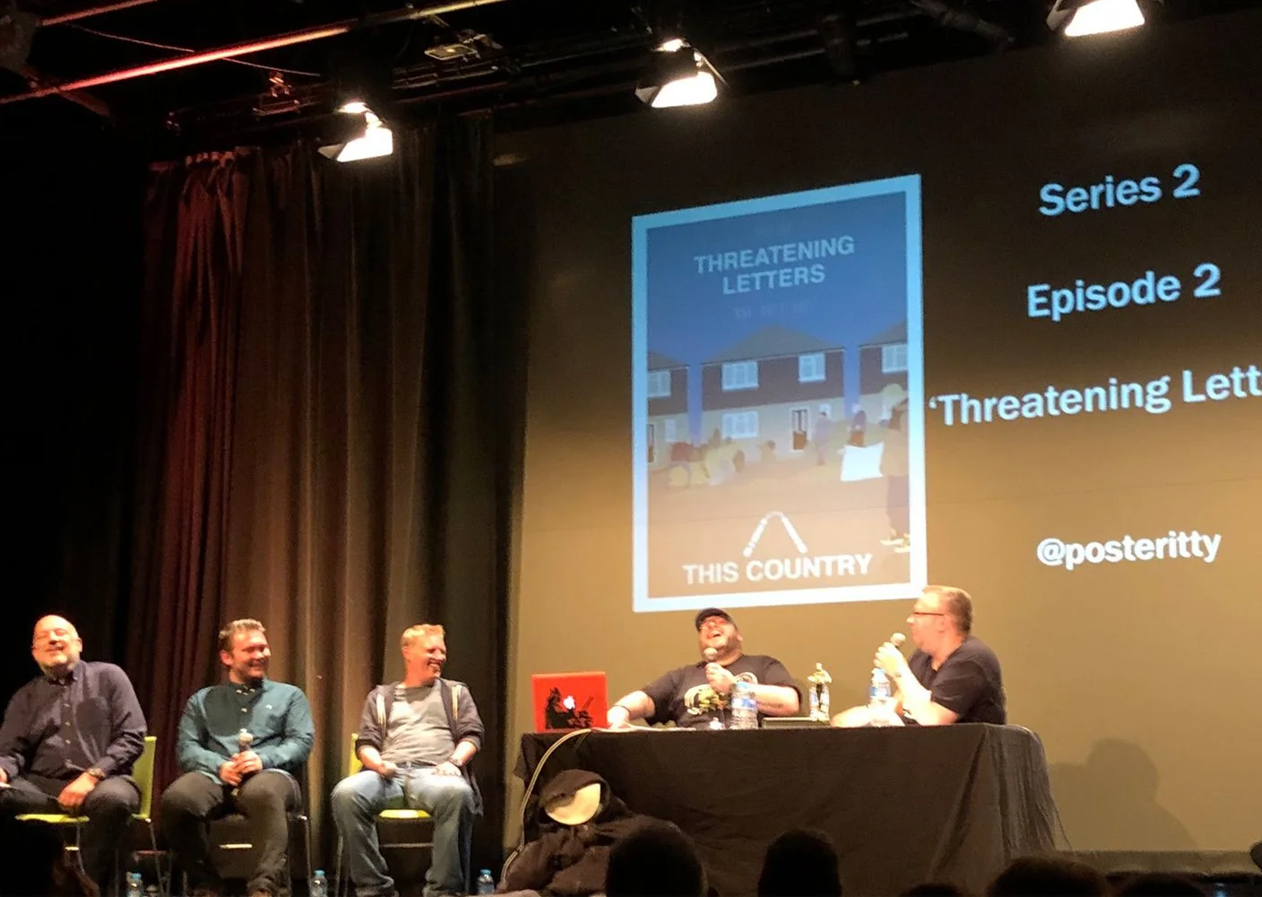 Four men seated on stage at a panel discussion, with a large screen behind them displaying a book cover titled 'Threatening Letters' and some text about series and episode numbers.