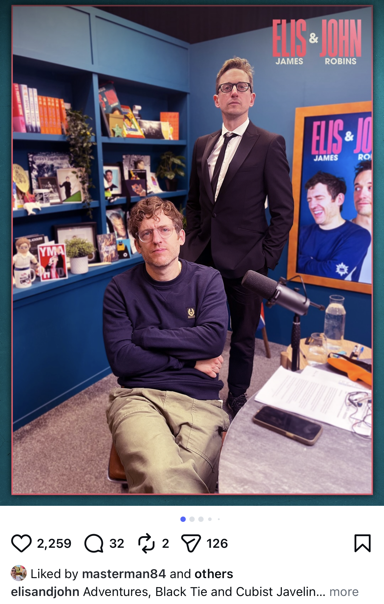 Two men in an office or studio setting, one sitting in front of a table with paperwork and a smartphone, the other standing behind him in a black suit and glasses, with bookshelves and a large poster of the same two men in the background.