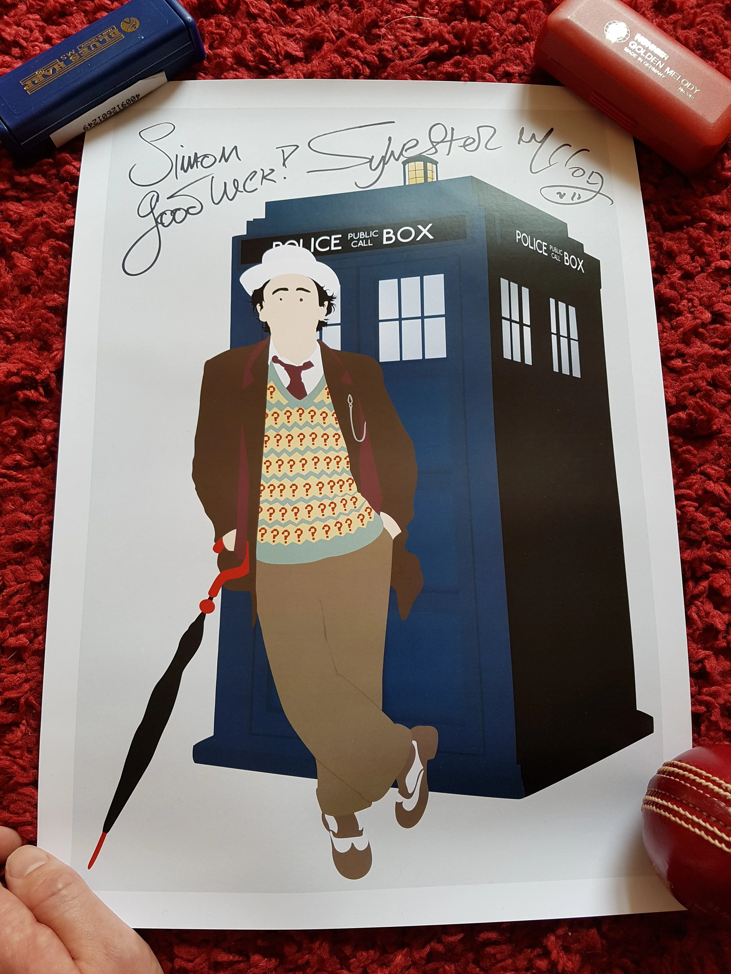 A printed illustration featuring the TARDIS from Doctor Who, with a character wearing a colorful vest with question marks, brown jacket, white hat, and beige pants standing in front, on a white background. Handwritten text above says, "Simon? Good lu