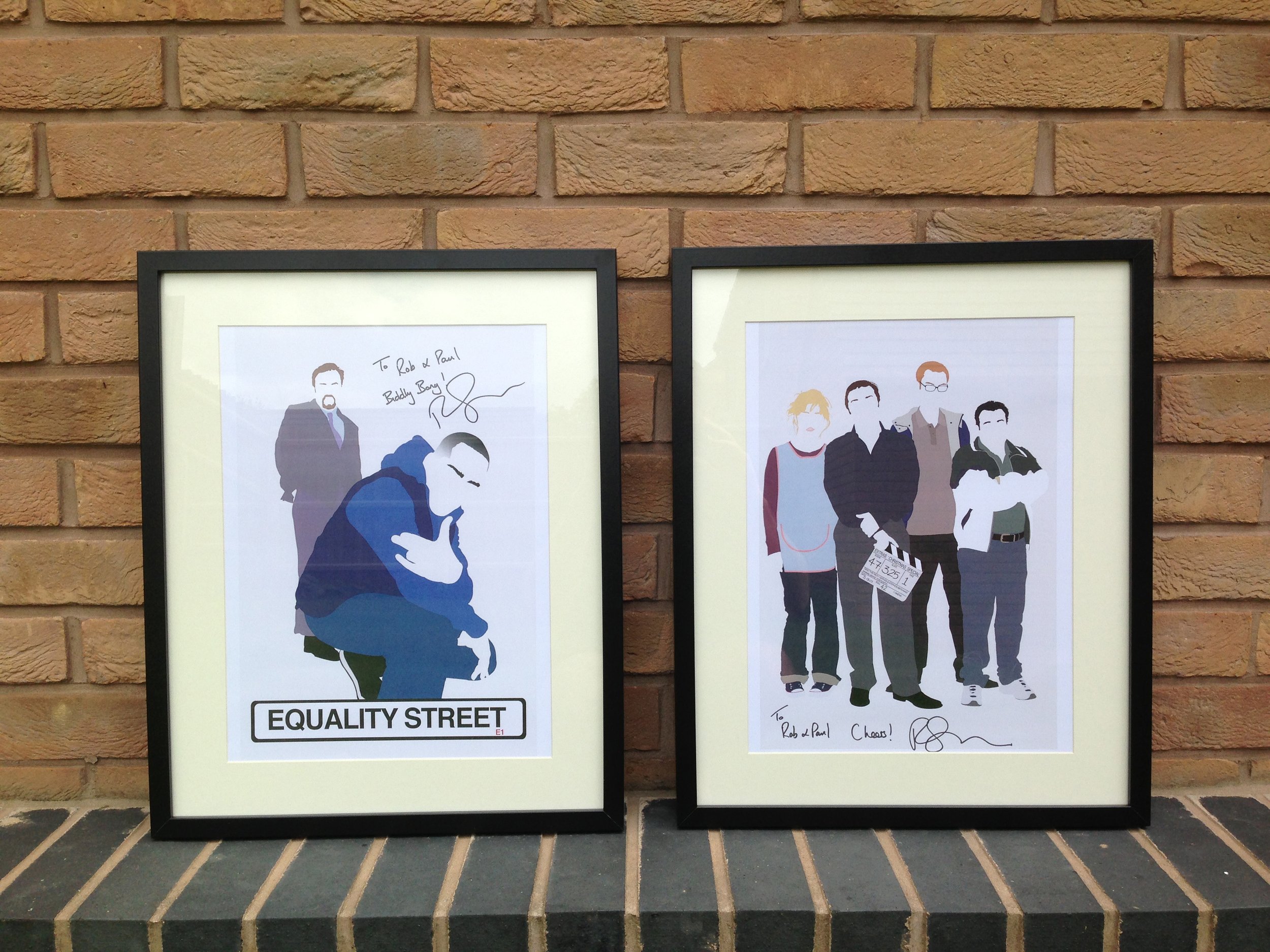 Two framed posters on a brick wall. The left poster features illustrations of diverse people with the text "Follow Your Street" and "Equality Street." The right poster shows a group of four people with signatures and the message "To Rob & Paul, Cheer