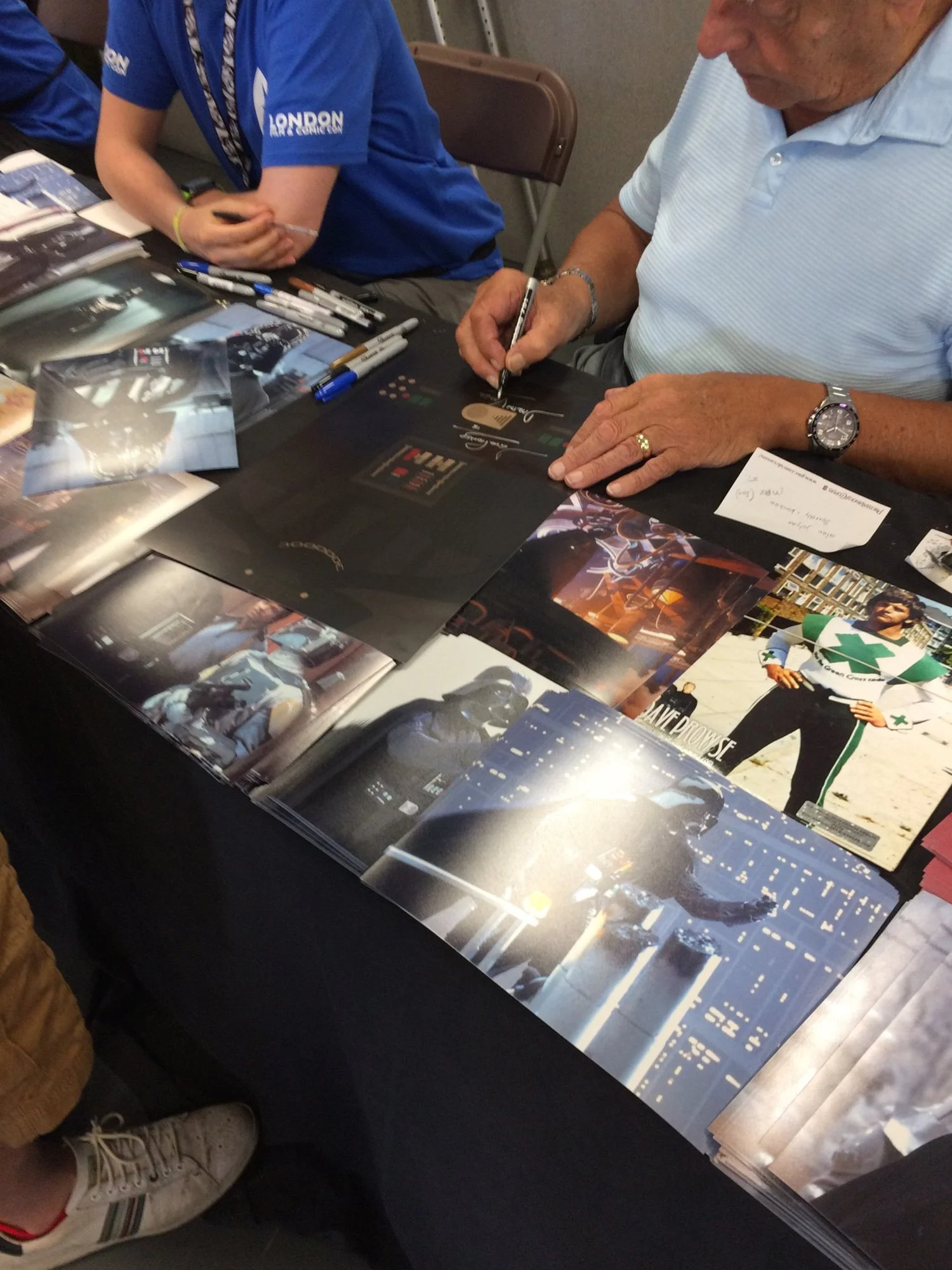 Several glossy posters of cars and city scenes displayed on a table. Two people are signing autographs on the posters; one is an older man wearing a light blue polo shirt, a watch, and a ring. The other person is wearing a blue shirt with white text 