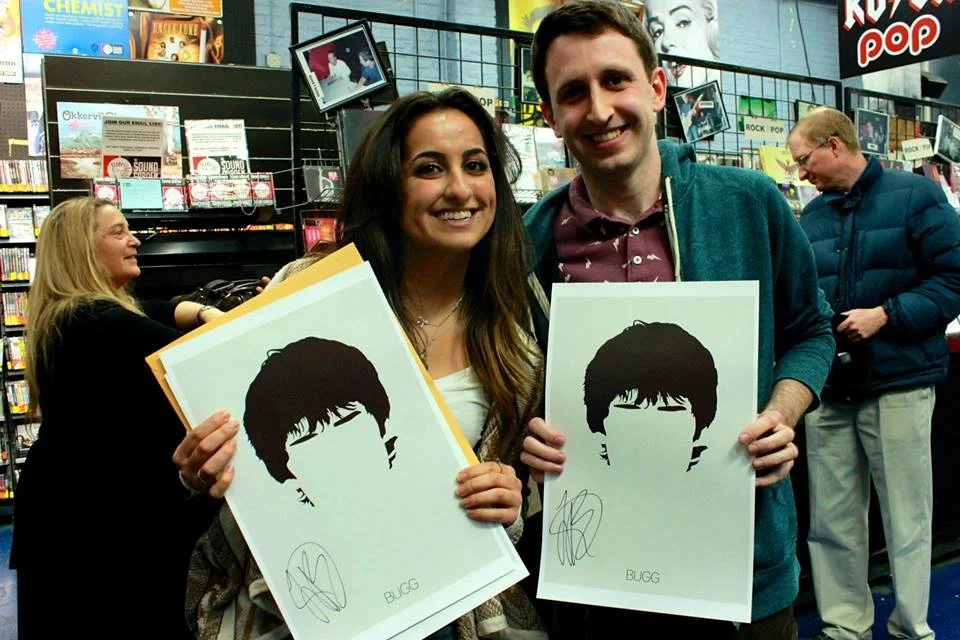 A woman and a man are holding signed posters of a stylized black-and-white portrait of a person's hair and face, smiling inside a bookstore or music store.