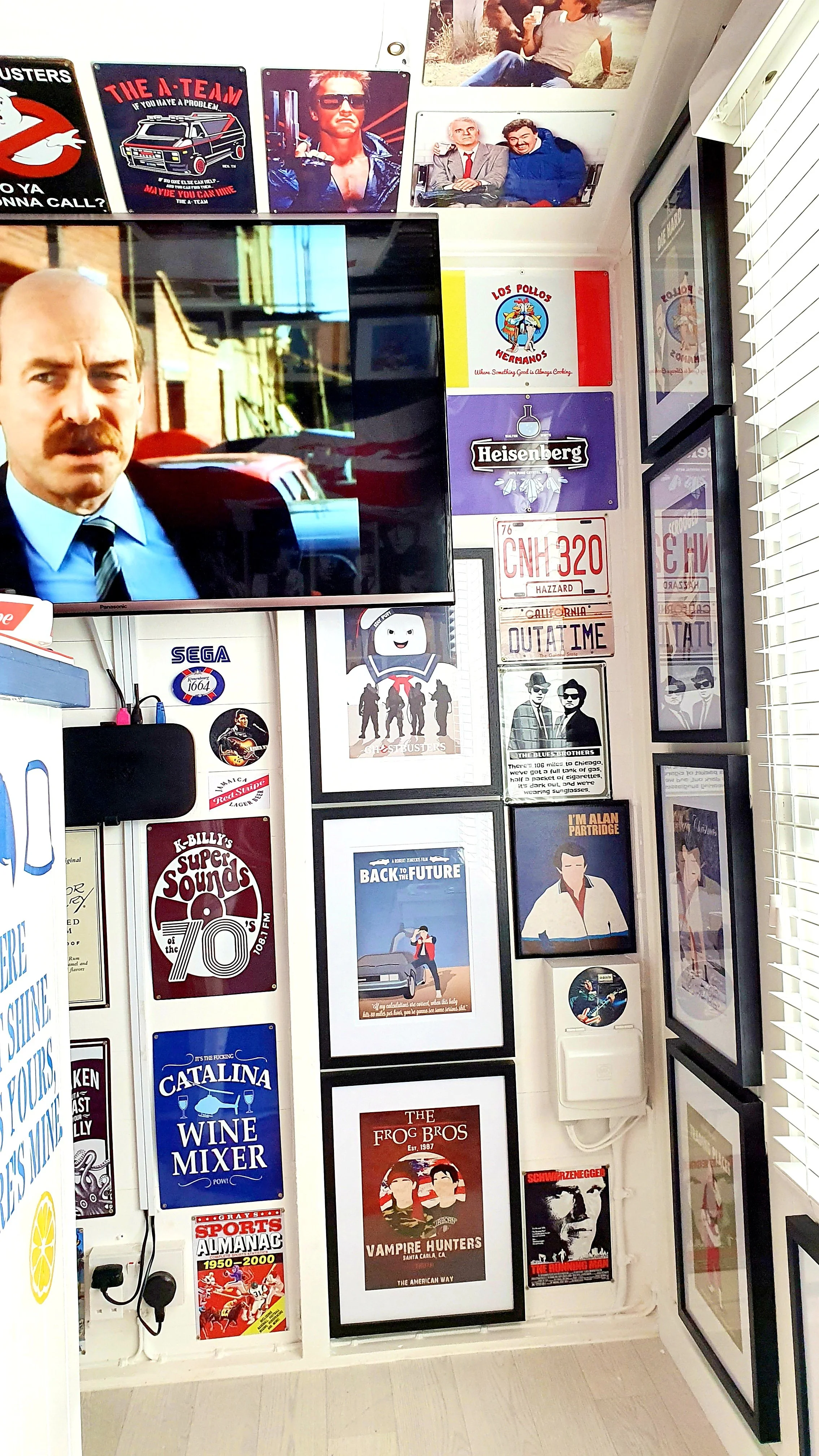 A wall decorated with various posters and framed pictures, including a large TV screen displaying a scene from a movie or TV show. Posters feature movies like 'Back to the Future', 'Vampire Hunters', and a 'Super Sounds of the 70s' album cover. There