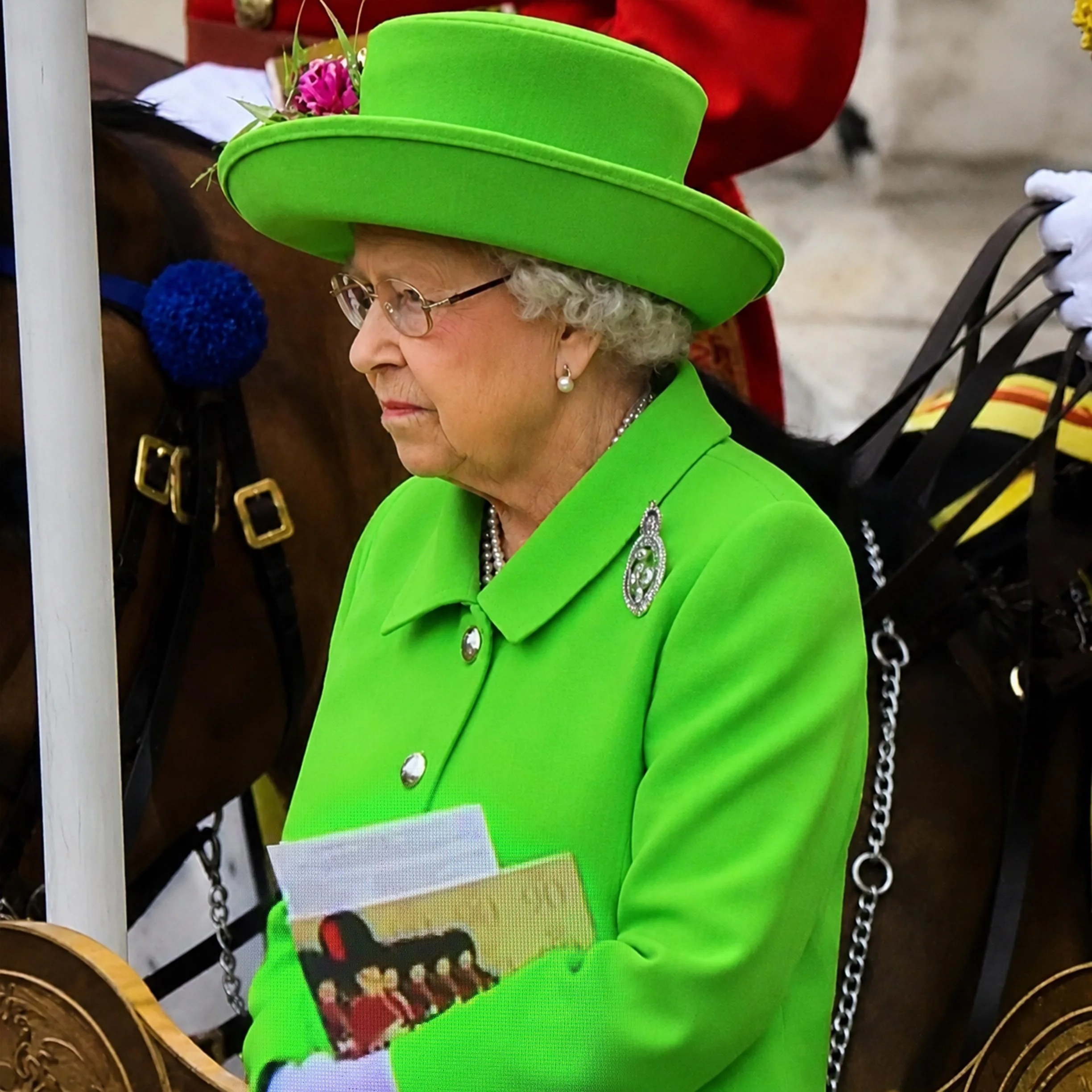 A woman dressed in bright green attire, including a hat decorated with pink flowers, is seen in profile with white curly hair and pearl earrings.
