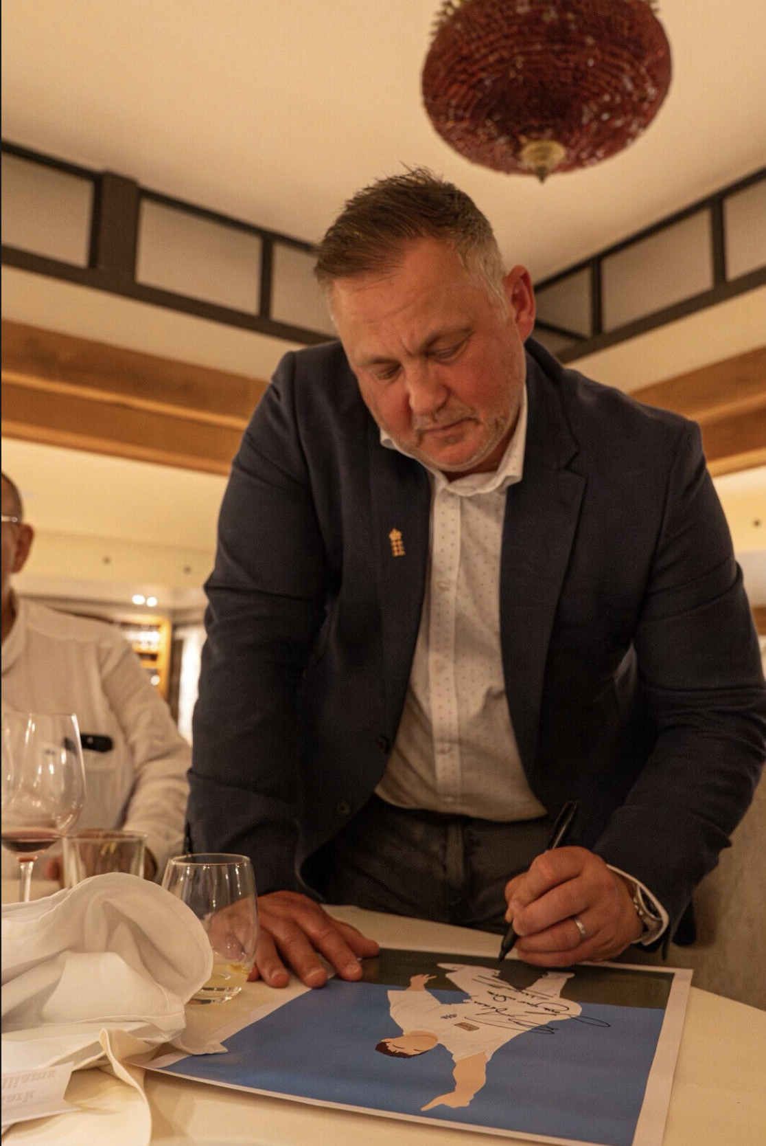 A man in a suit signing a poster at a dinner event, with wine glasses and plates on the table.