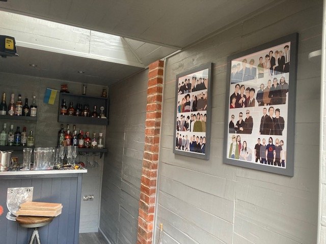 A bar area with bottles of liquor on shelves, glasses hanging upside down, and framed group photos on the wall.