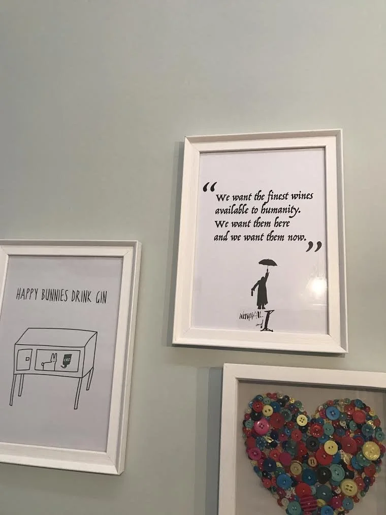 Three framed pictures on a light-colored wall: one with a quote about fine wines, one with an illustration of a bunny with the text "HAPPY BUNNIES DRINK GIN," and one with a heart shape made of colorful buttons.