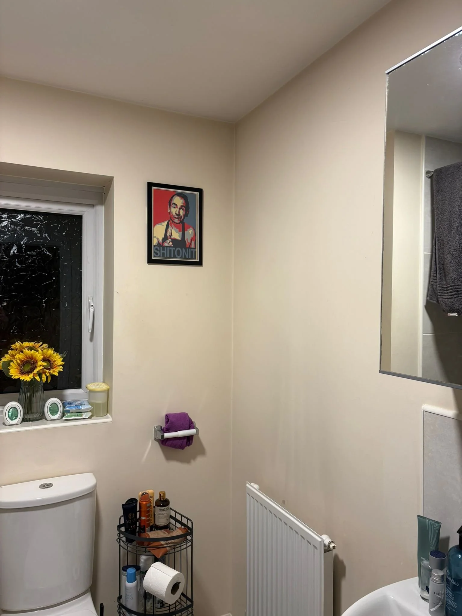 A small bathroom with cream-colored walls, a window with a sunflower bouquet, and a framed portrait of a person with the word 'SHITONI' underneath. The bathroom features a white toilet, a black metal storage rack with toiletries, a wall-mounted mirro