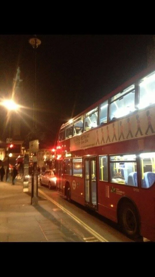 Nighttime street scene with a red double-decker bus, cars, and pedestrians, illuminated by streetlights.