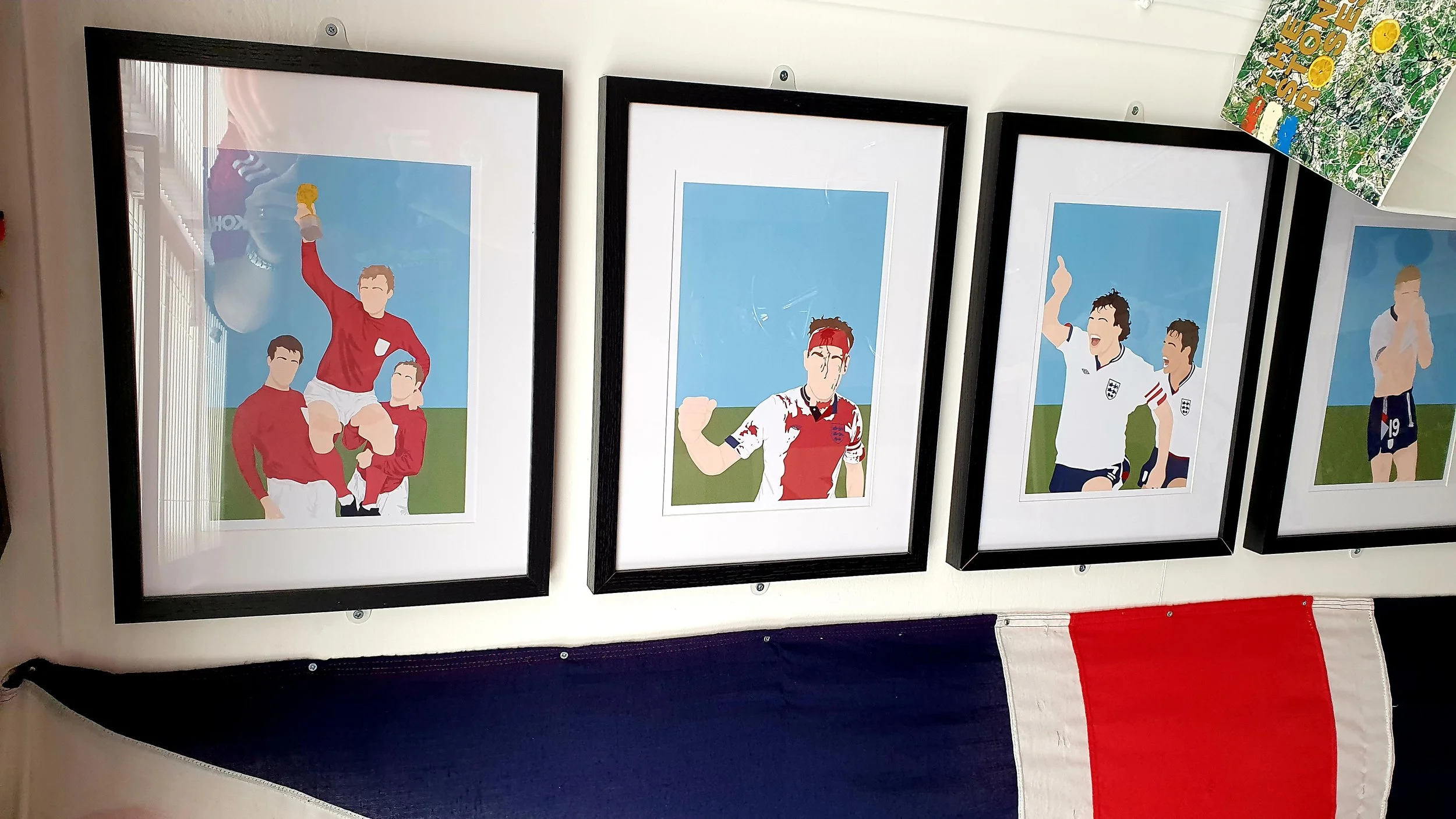 Set of four framed sports illustrations hung on a white wall, depicting soccer players celebrating, with a flag with blue, white, and red stripes hanging below.