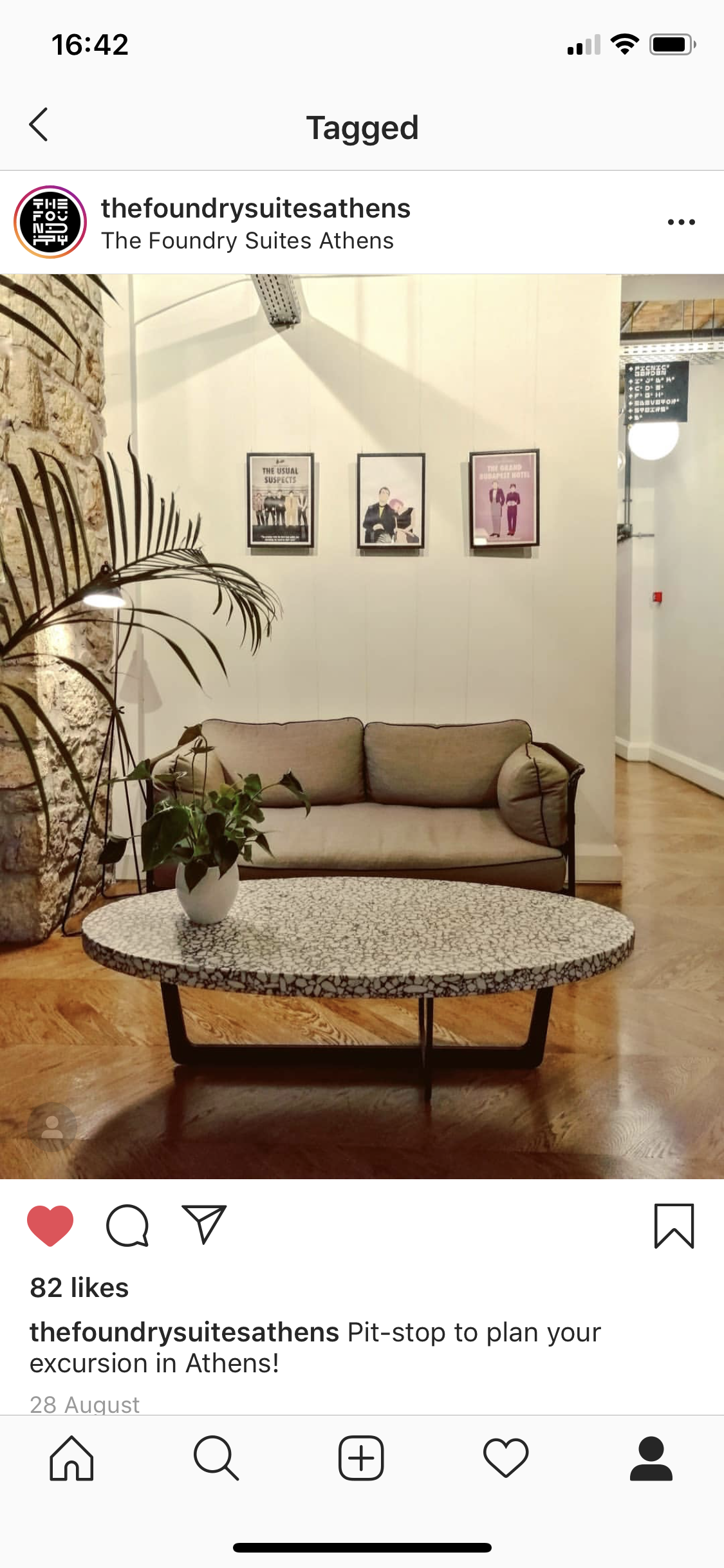 Interior of a hotel lobby with a beige sofa, a round coffee table with a white vase and green plant, a stone wall on the left, framed posters on the wall, and wooden flooring.