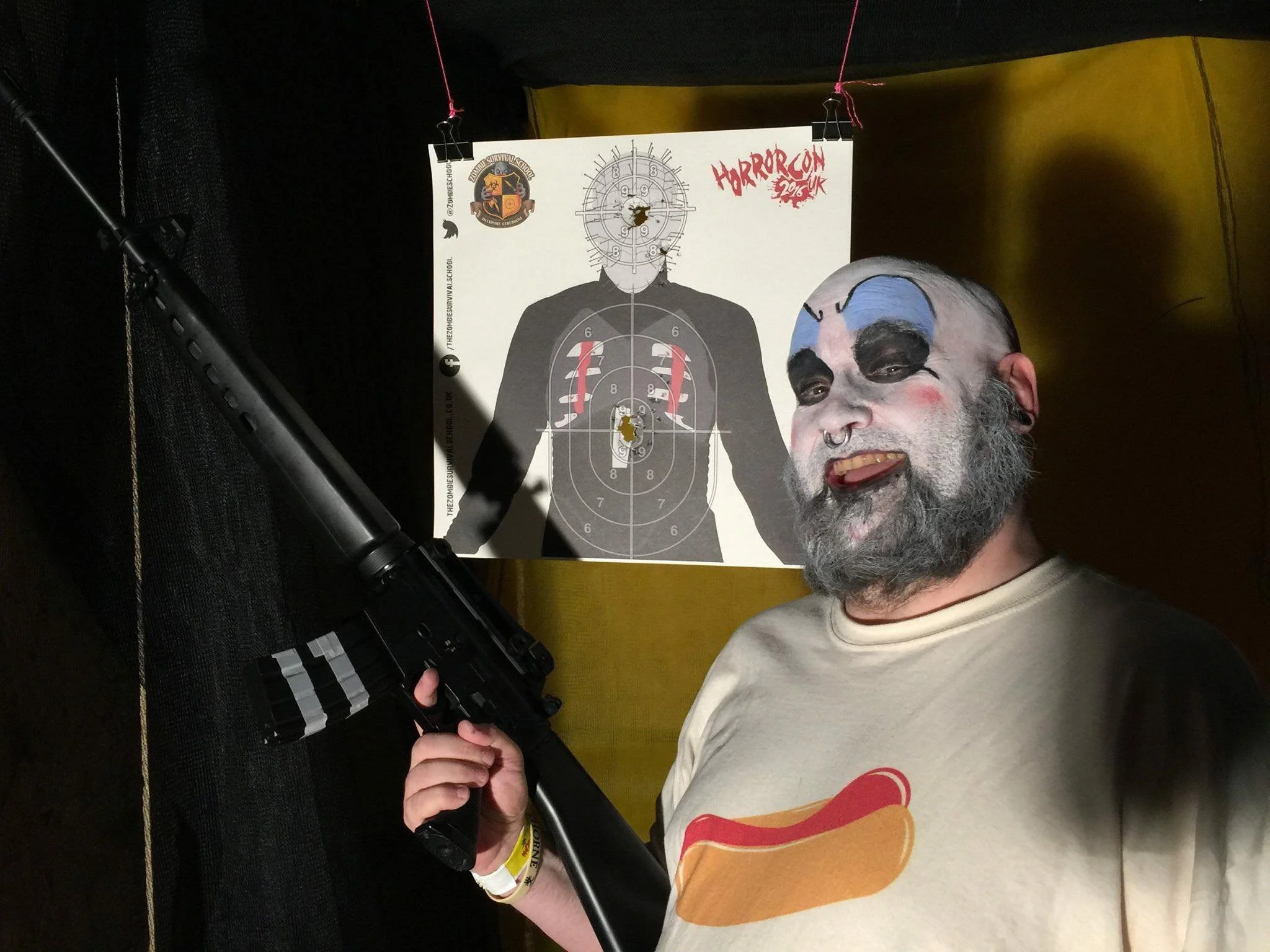 Person with clown makeup and a beard, wearing a beige shirt with a hamburger graphic, holding a black firearm, standing in front of a horror-themed poster with a person's silhouette and target design, at an event named 'HorrorCon 2016 UK'.